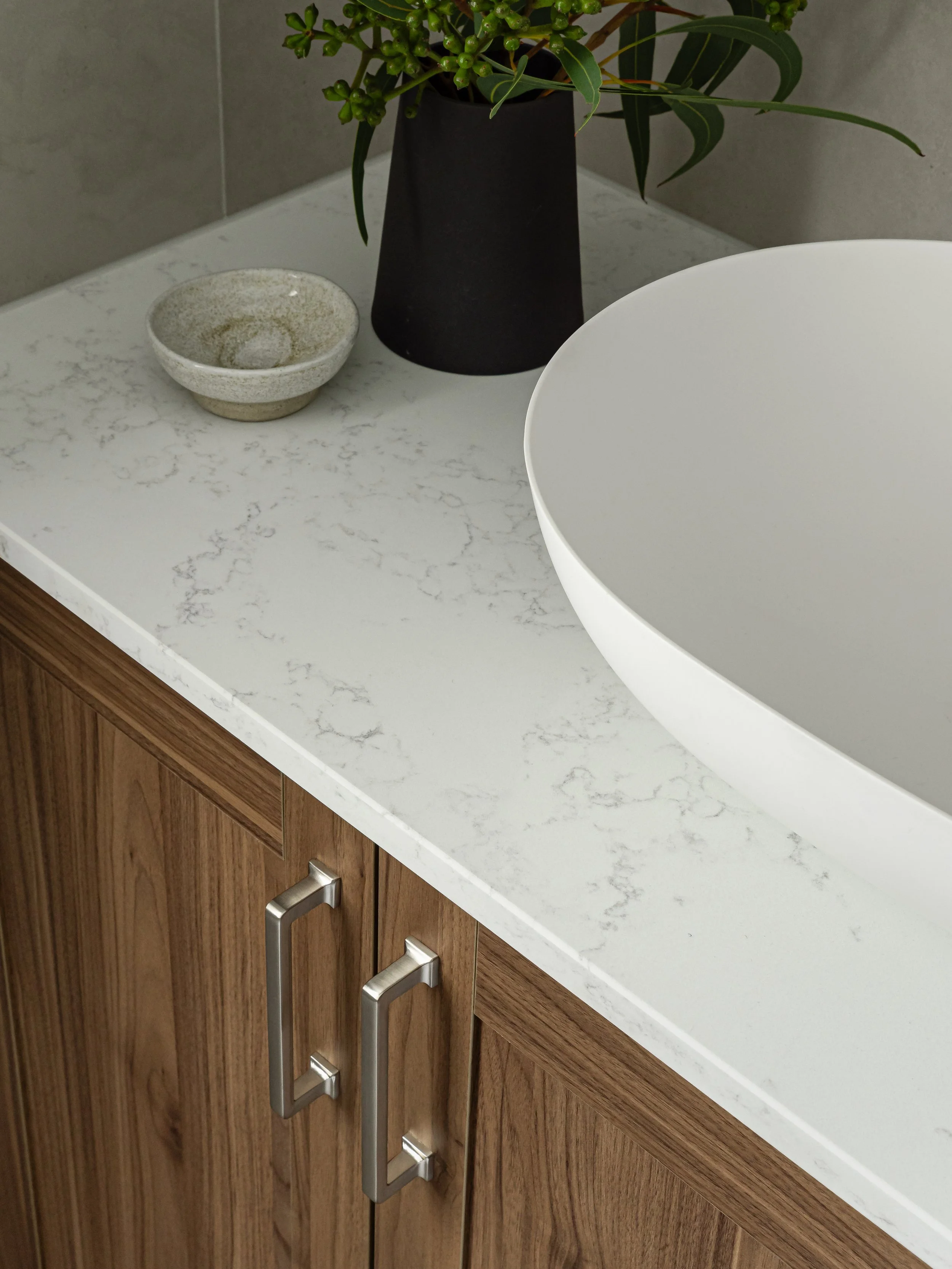 angled photo of bathroom vanity, with white stone top and medium wood cabinet, on top is a vase with leaves 