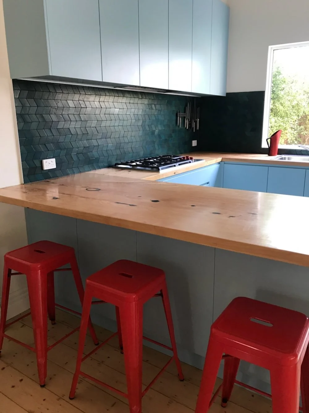 Kitchen renovation, with refined, practical, and balanced choices. Shows wooden tops and floor, blue cabinets, dark green tiles, red metal stools.