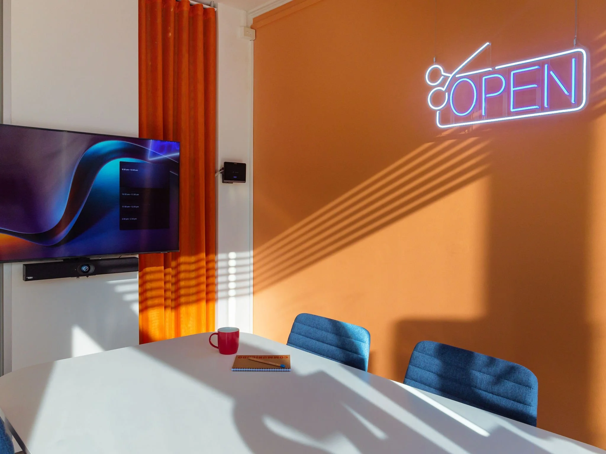 View of the meeting room, showing a TV screen, table, and chairs. In the background on the orange wall is a neon open sign.