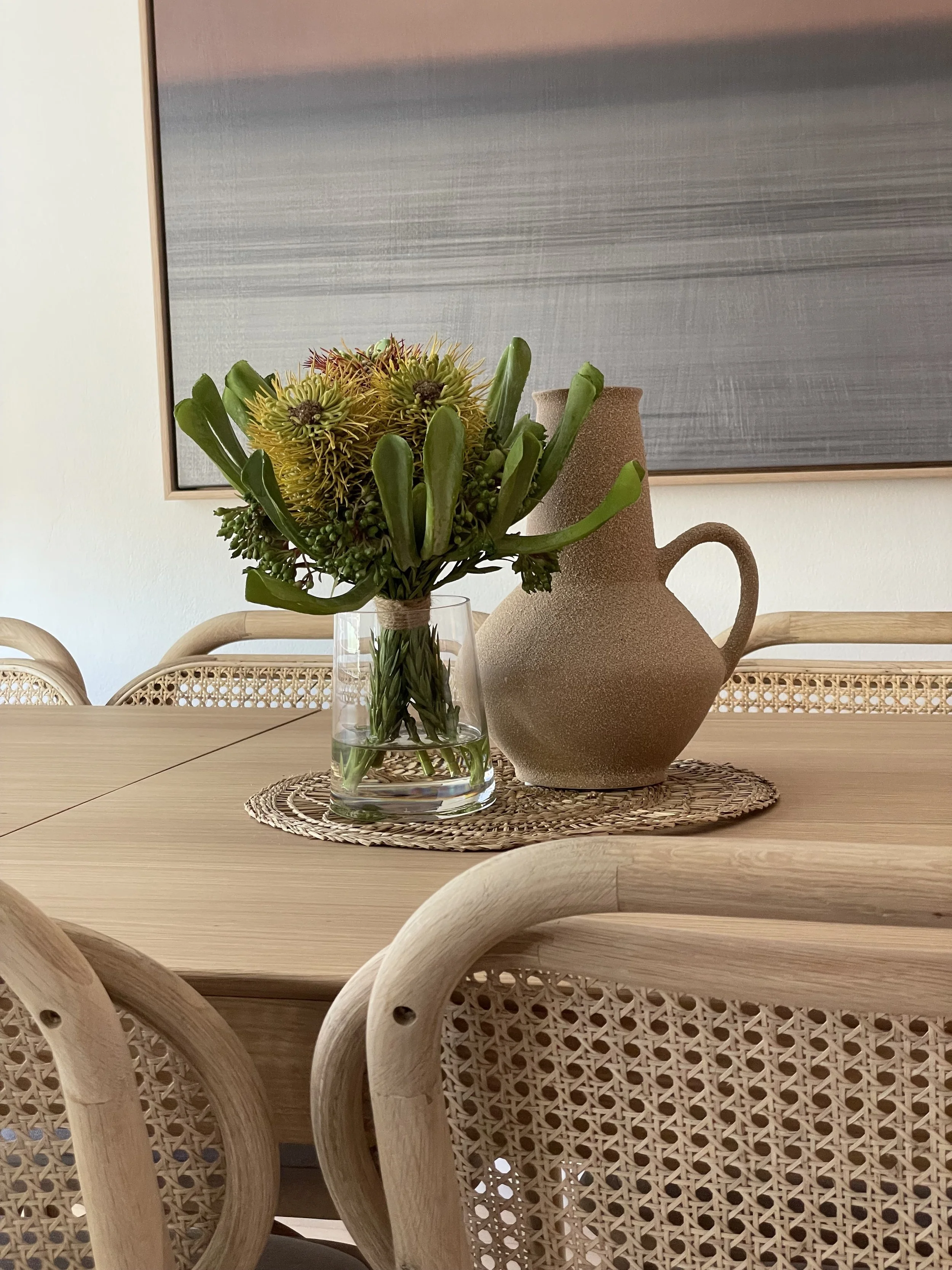 A wooden dining table with a woven placemat, a glass vase with yellow-green flowers and greenery, and a textured ceramic pitcher. In the background is a large abstract painting with horizontal gray and pinkish tones.