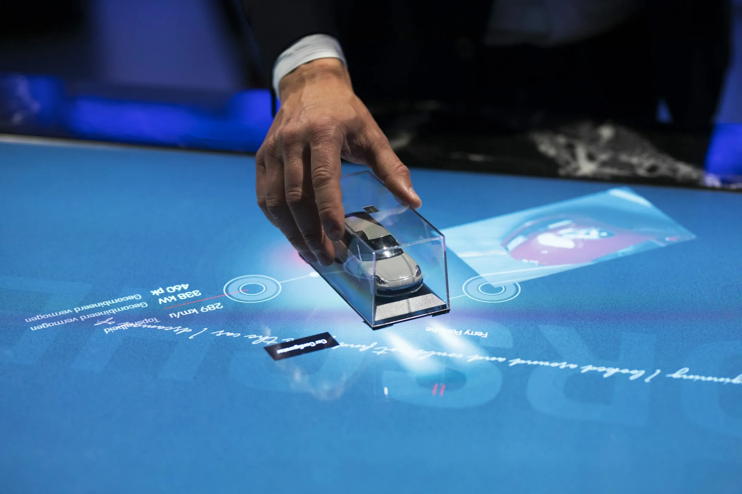 Una mano tocando una maqueta de automóvil en una mesa interactiva digital con gráficos y texto en el fondo oscuro.