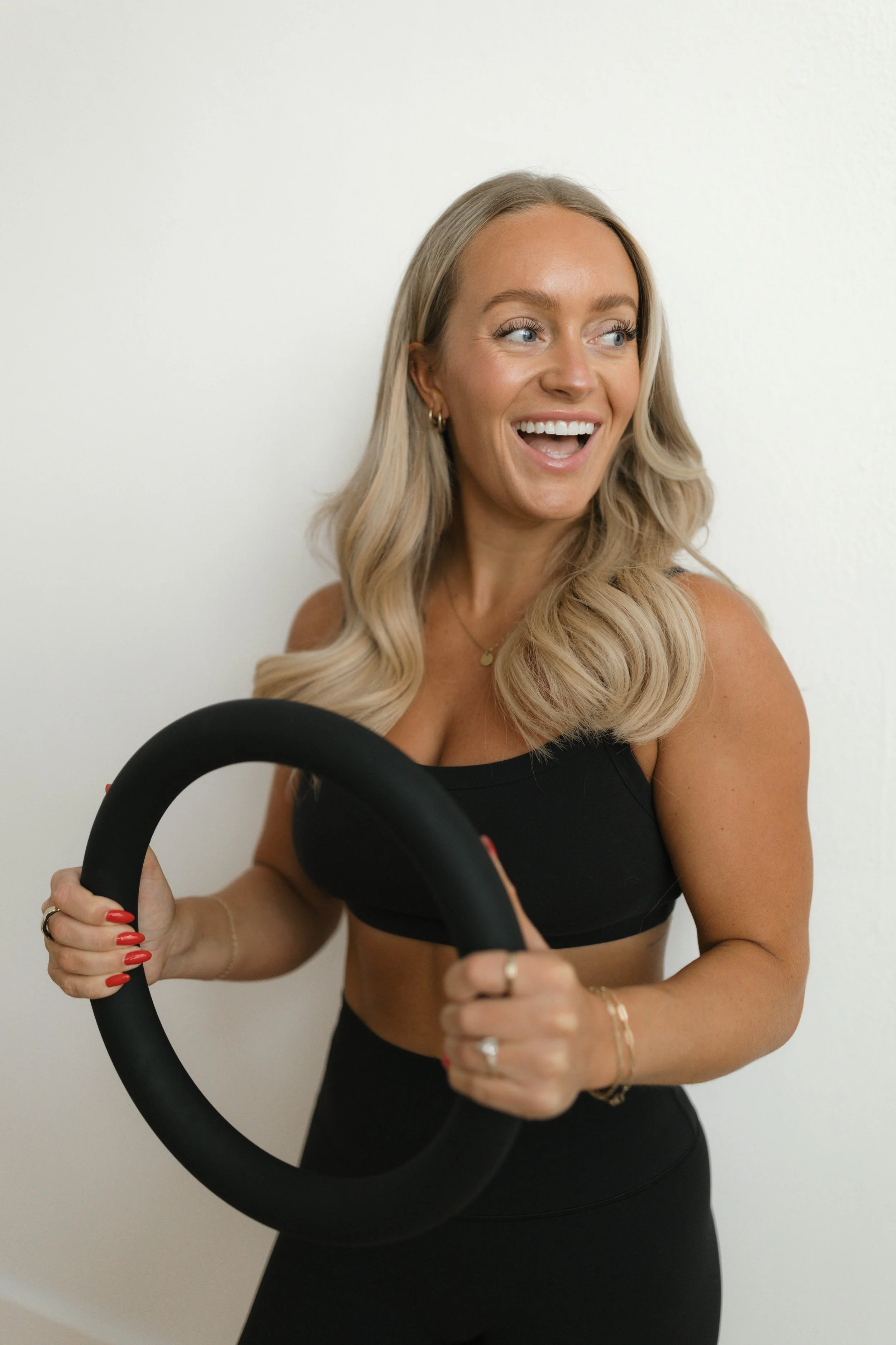 A woman with long, wavy blonde hair, wearing a black sports bra, holding a large black exercise ring, and smiling while looking to the right, against a plain white wall background.