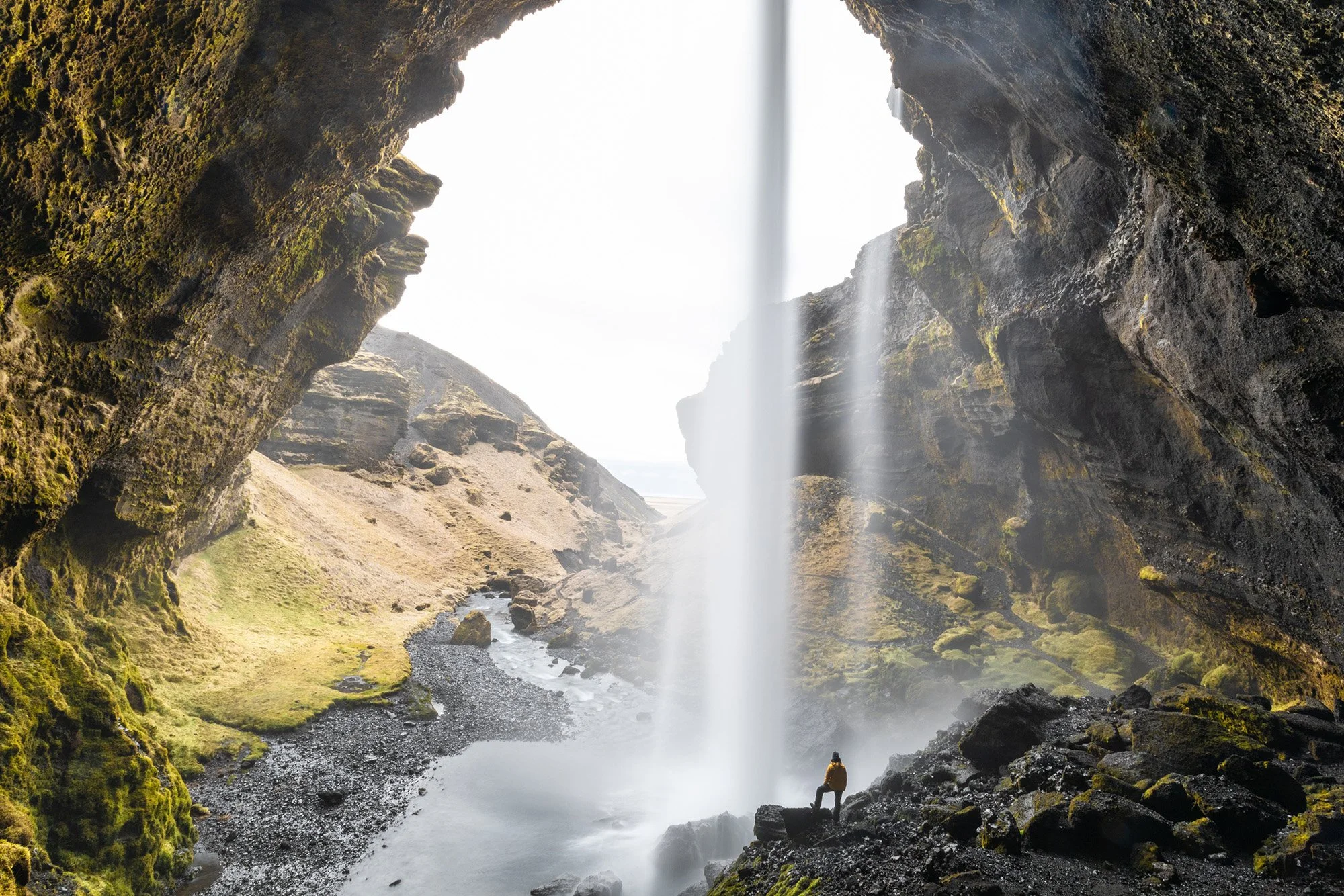 Eine Person steht vor einem Wasserfall in einer Höhle mit grünen, moosbedeckten Felsen, Blick nach draußen auf die Landschaft.
