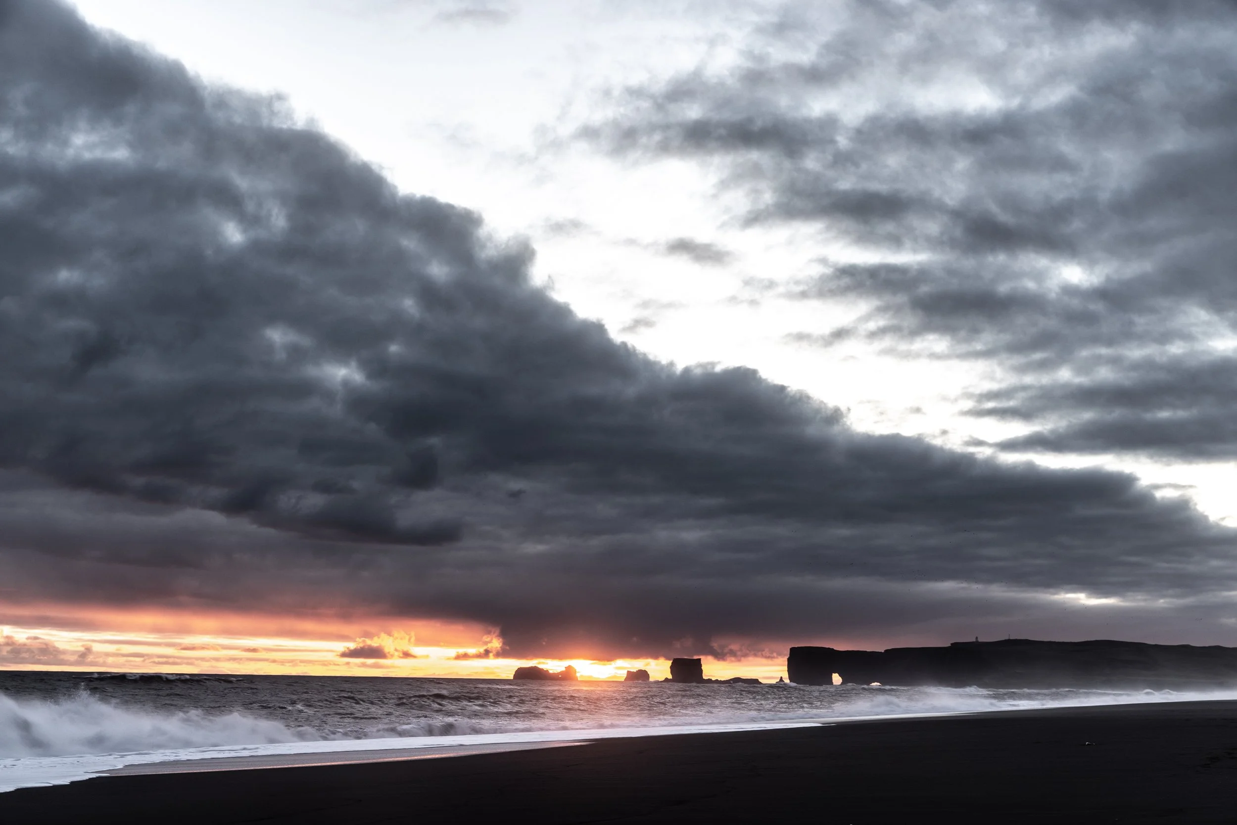 Ein dunkler Strand bei Sonnenuntergang mit Wolken am Himmel und Felsen im Wasser.