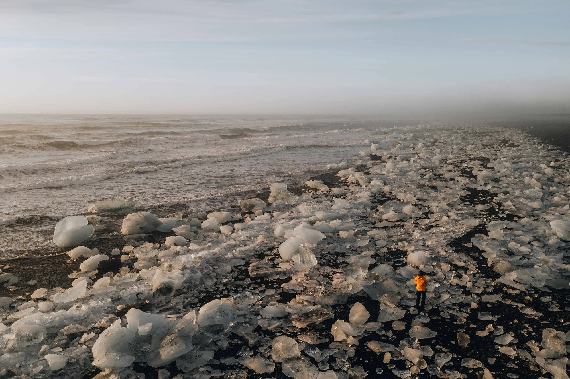 Ein Mensch in orange warmer Jacke steht auf einem dunklen, steinigen Strand, der mit großen Eisstücken übersät ist. Im Hintergrund sind Wellen im Meer sichtbar, und der Himmel ist bewölkt.