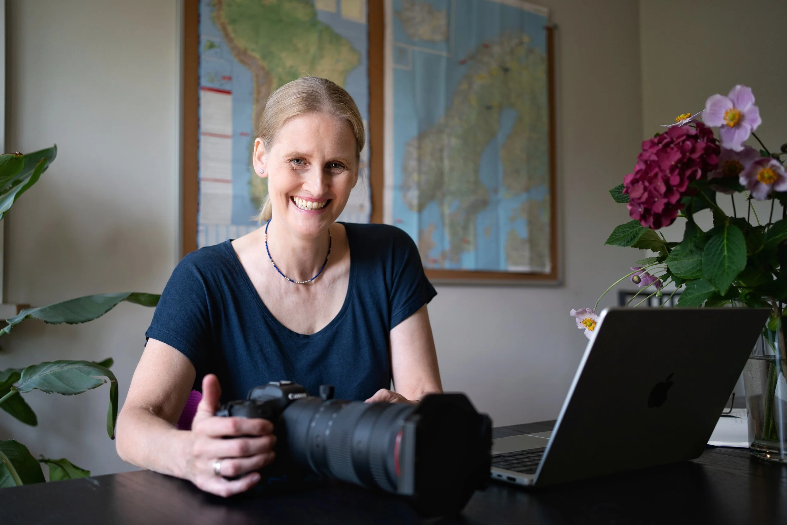 Eine lachende Frau sitzt an einem Tisch mit Kamera, Laptop und Blumen vor einem Wandbild mit einer Karte.