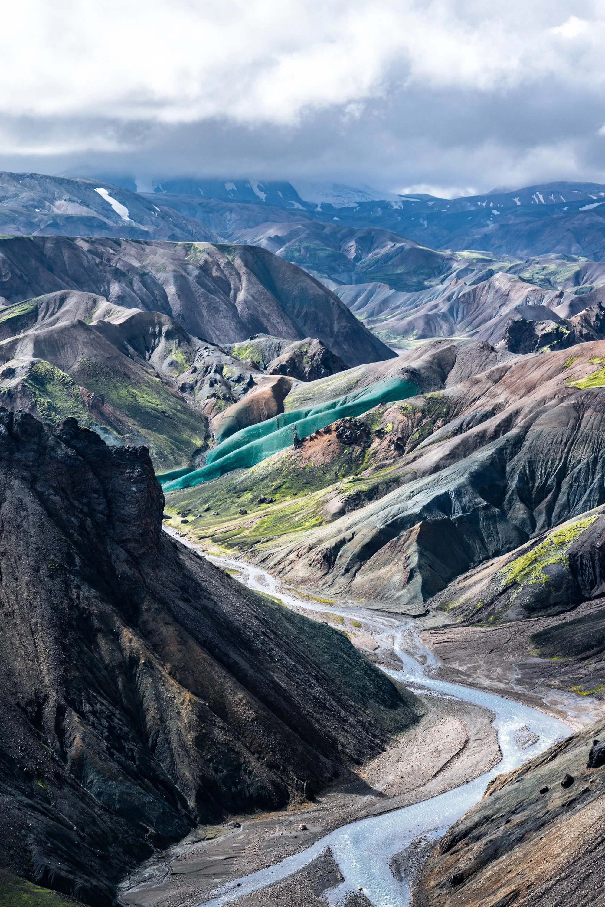 Berglandschaft mit einer schmalen, fließenden blauen Gletscherflüssigkeit in einem Tal, umgeben von bunten, zerklüfteten Bergen und bewölktem Himmel.