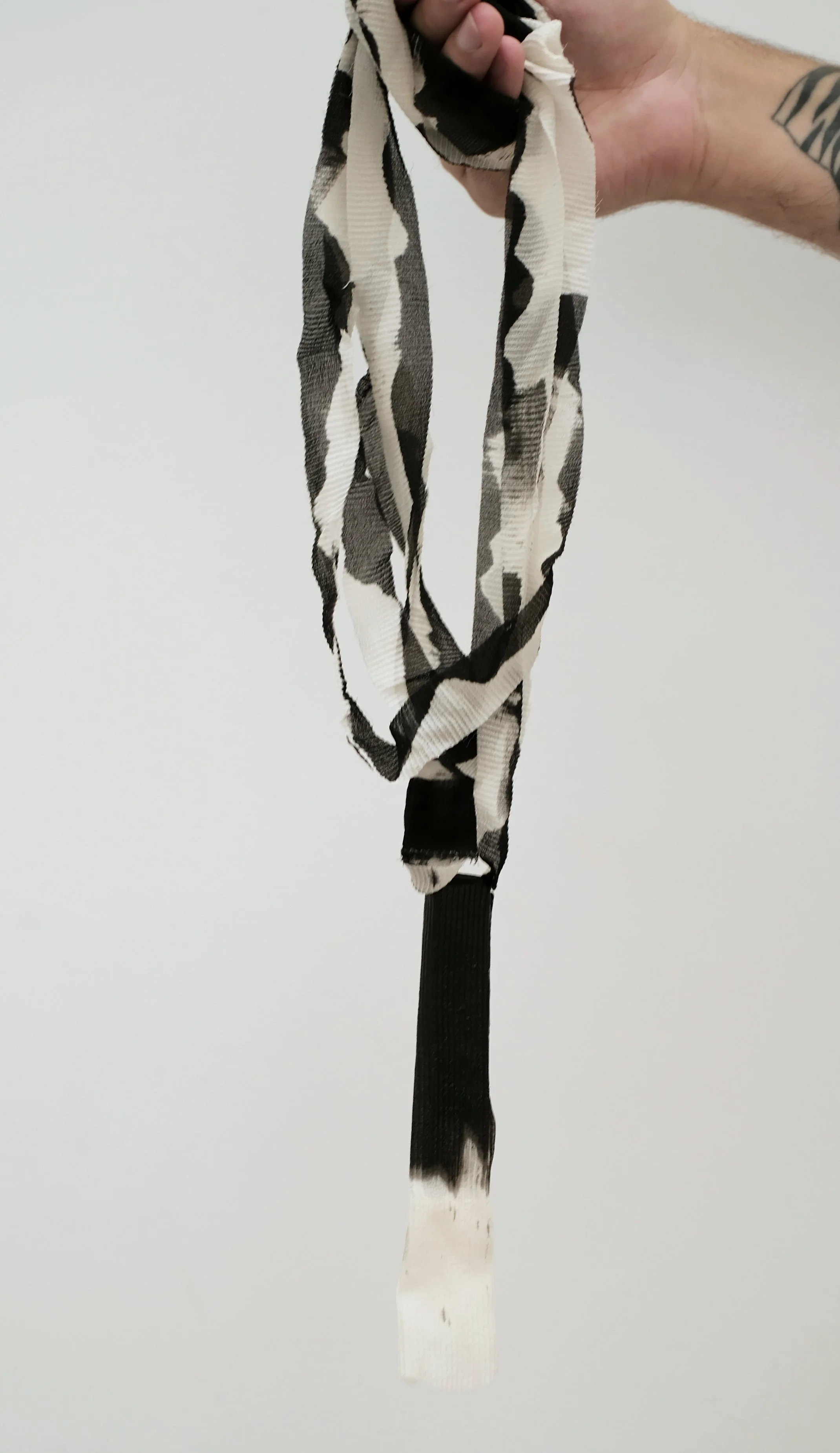 Une main tient un bandana noir et blanc avec un motif abstrait. La personne a une tatouage au poignet et porte un jean noir.