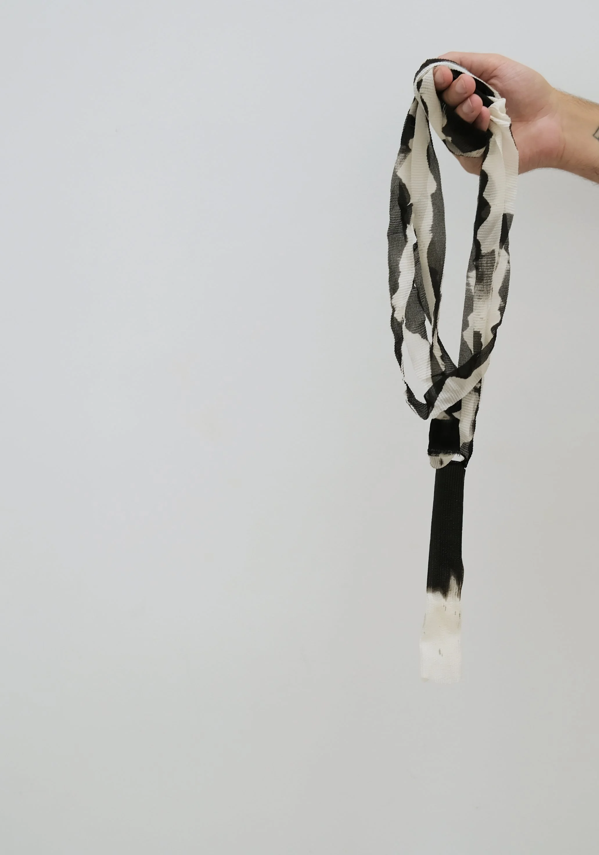 Une main tient un foulard en tissus noir, blanc et gris avec un motif à rayures et taches, suspendu à une bande noire, contre un fond blanc et simple.