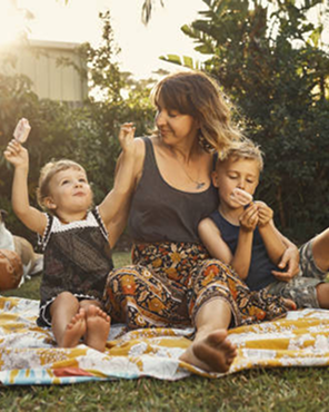 A woman sitting on a blanket outdoors with two children holding popsicles, surrounded by greenery.