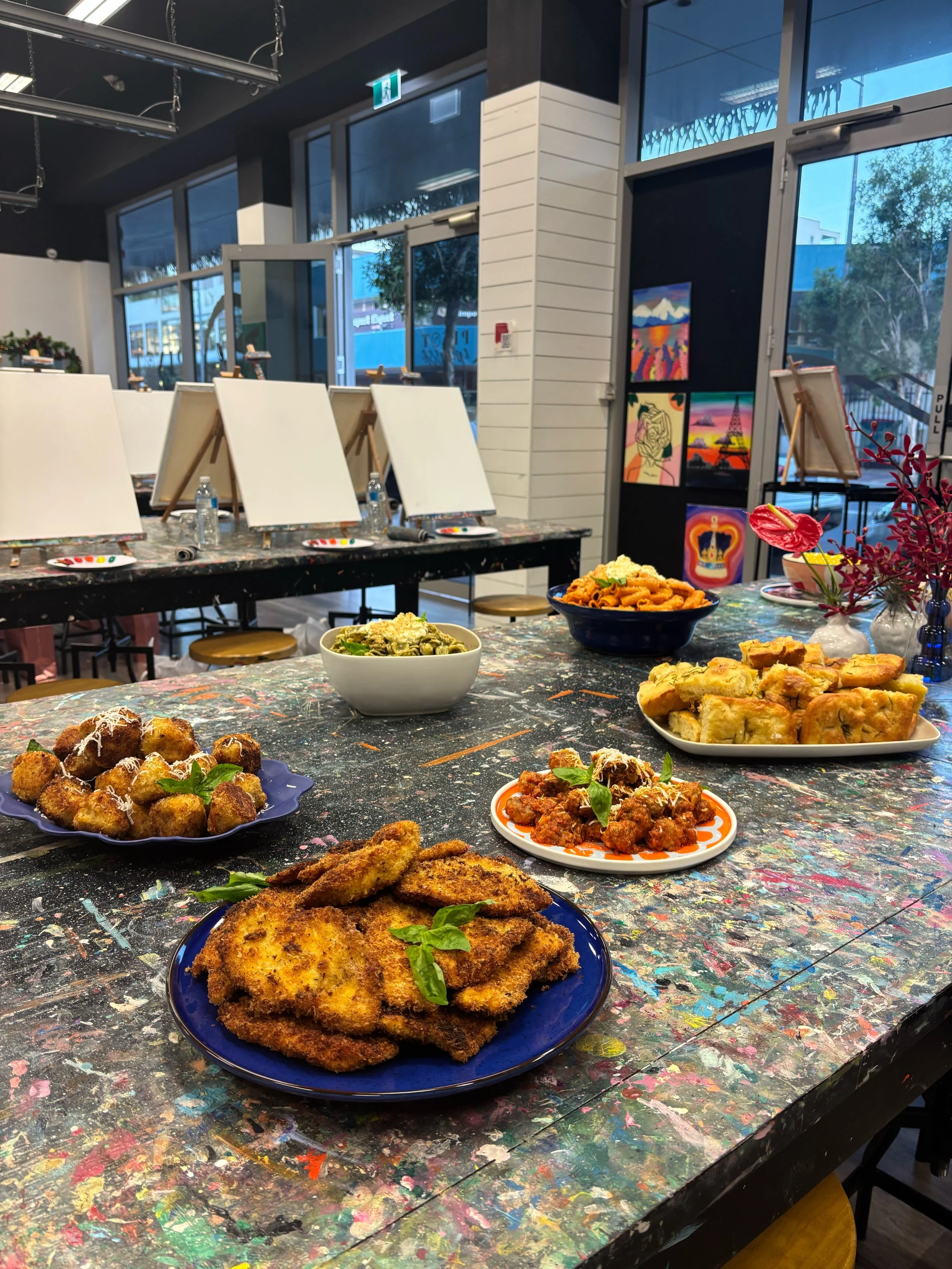 Variety of plates with fried foods and pasta on a colorful, artsy table in a restaurant or art studio space with canvases and artwork on the walls.