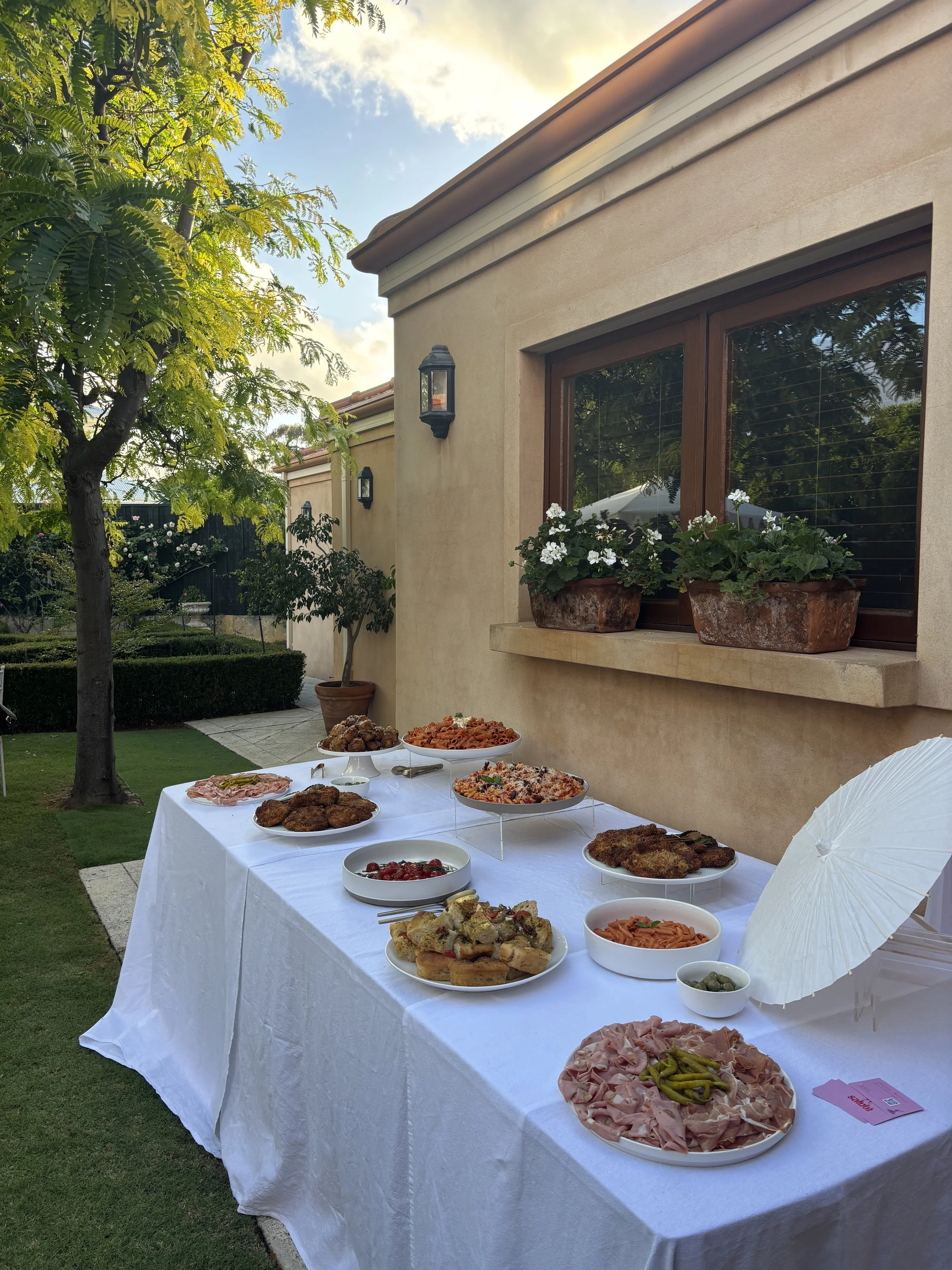 Outdoor buffet table with various dishes on a white tablecloth, set against a beige house wall with two flower boxes under a window, surrounded by green trees and grass.