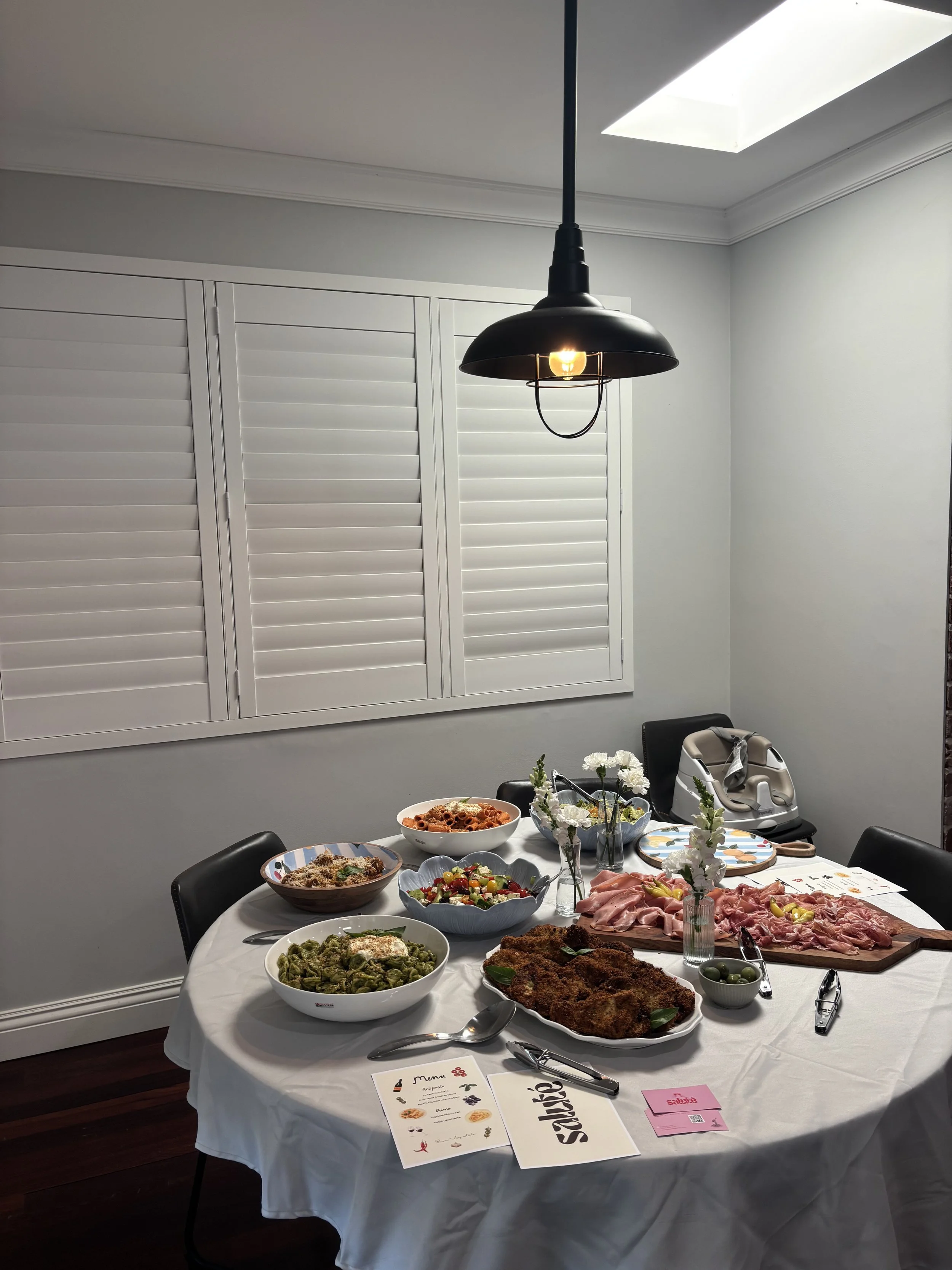 Dining table with various dishes including pasta, salad, breaded cutlet, and cured meats, decorated with flowers, in a room with a hanging black lamp and white window shutters.