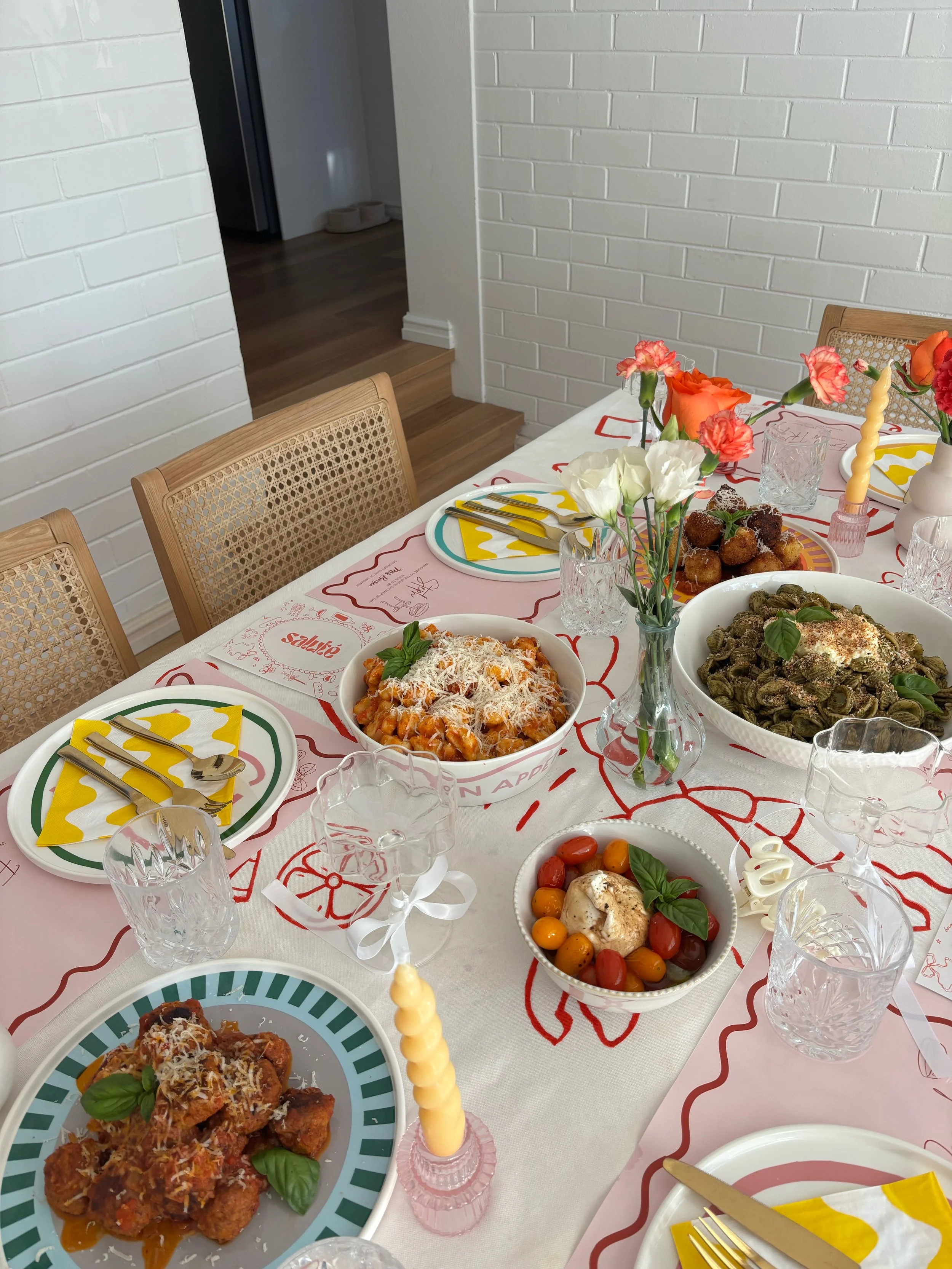 A festive dining table with various Italian dishes, floral centerpieces, candles, glasses, and colorful napkins with knives and forks, set in a bright room with white brick walls.