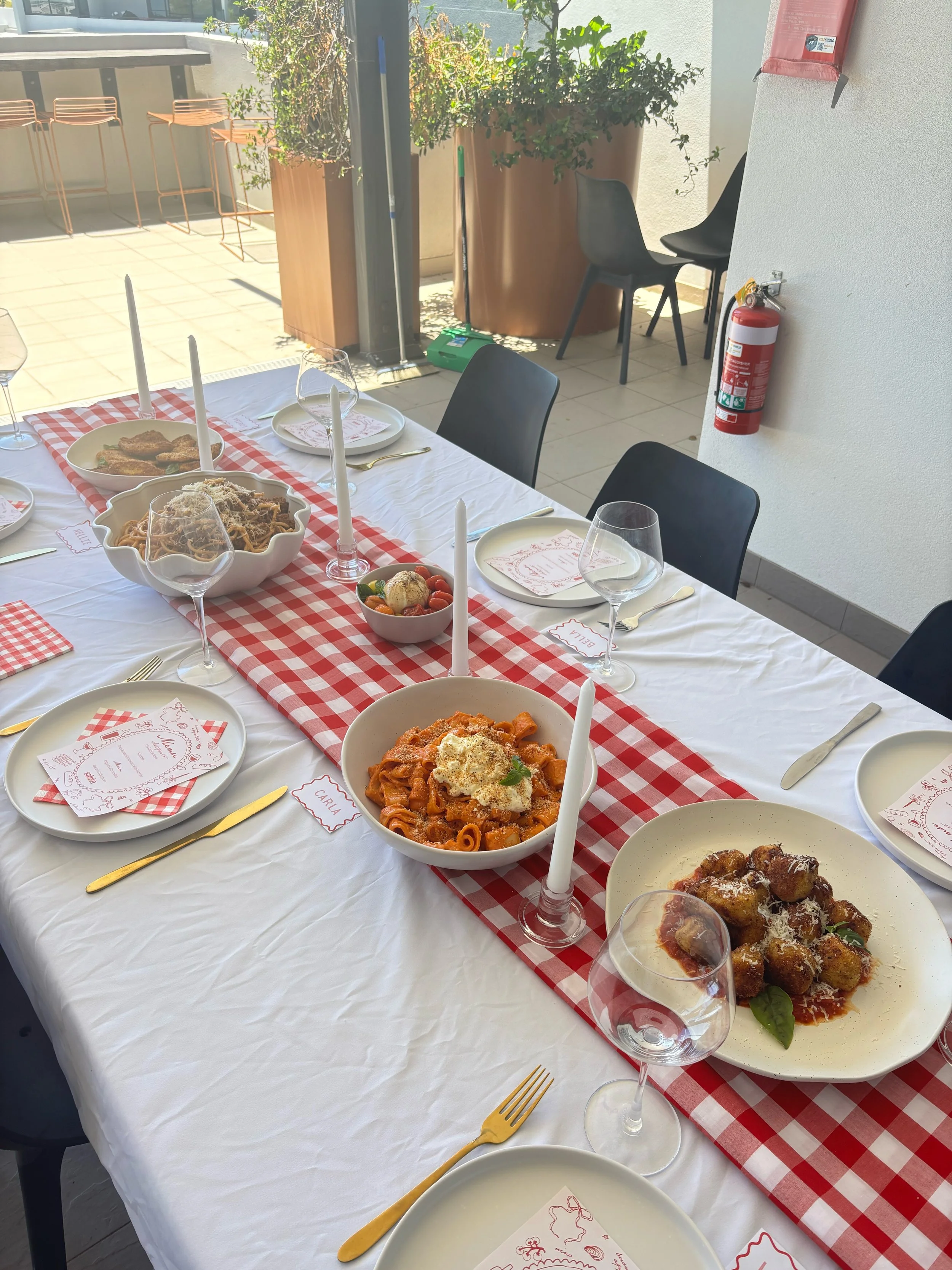 Table set for a meal with bowls of pasta, meatballs, and other dishes, surrounded by plates, wine glasses, and gold-toned utensils, with a red and white checkered table runner and place cards.