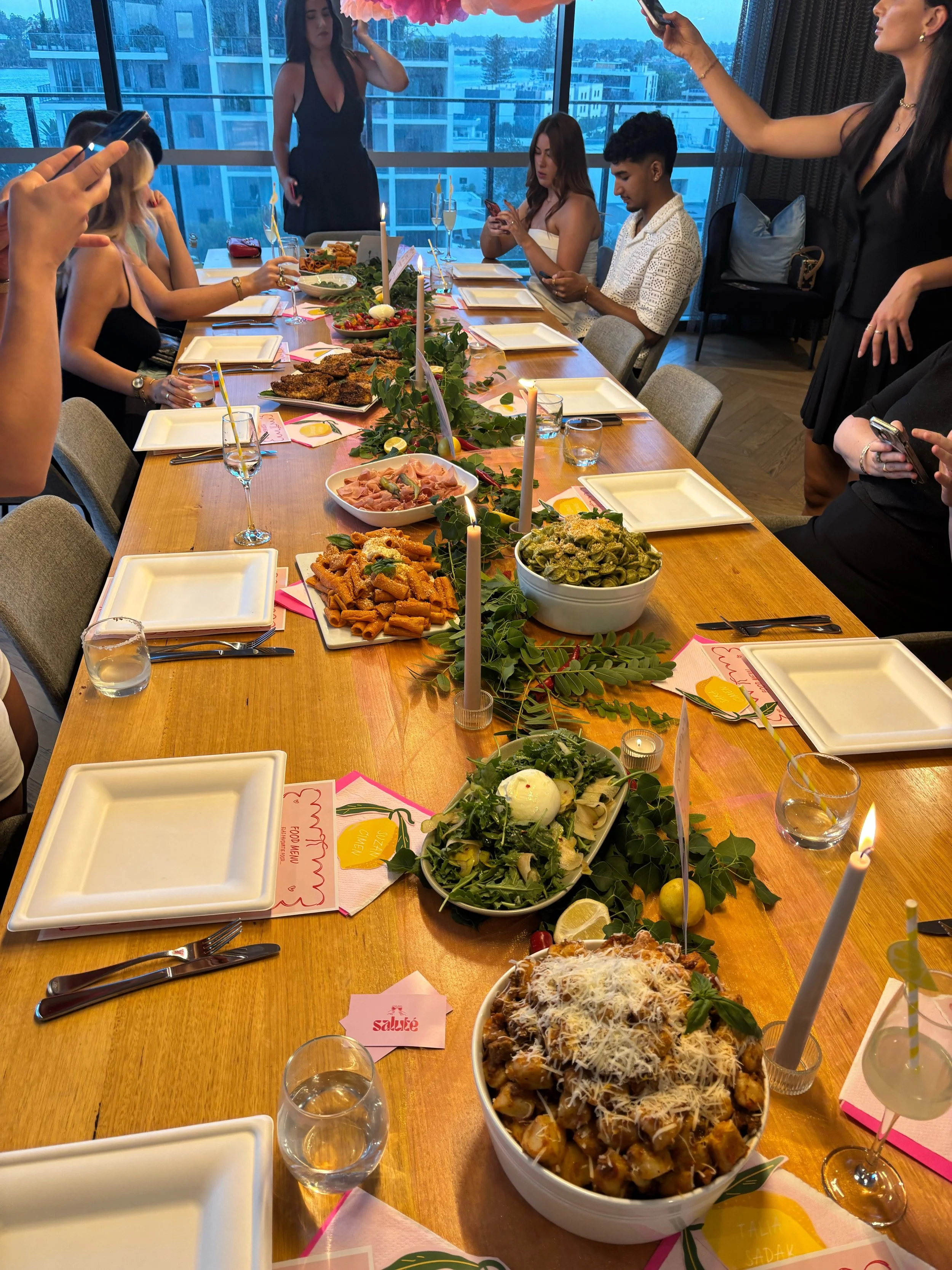 A long dining table set for a celebration with various dishes, green leafy decorations, lit candles, and pink and yellow place cards. People are gathered around, some taking photos, with a cityscape visible through large windows in the background.