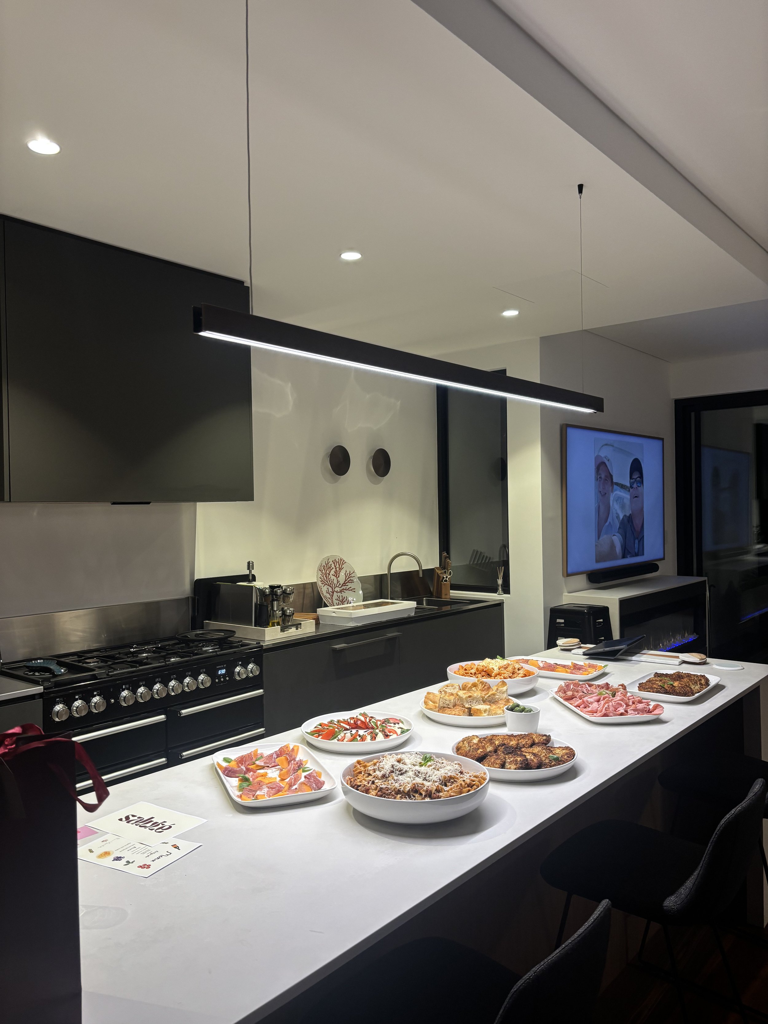 Kitchen with black cabinets and a white countertop displaying various dishes including salads, bread, and cooked meats. A television shows two people and the room has modern lighting and decor.