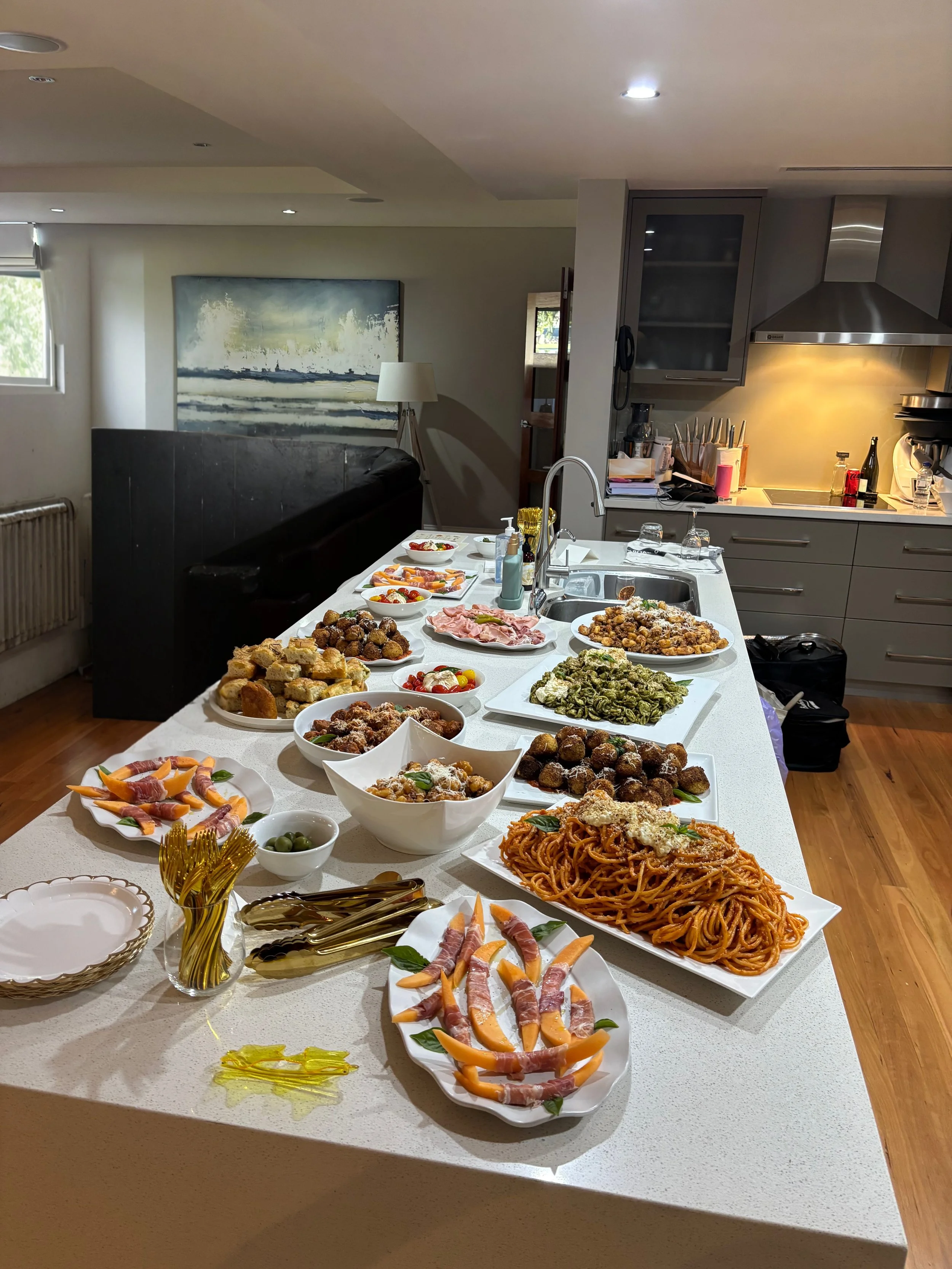 A kitchen island with various dishes including spaghetti, meatballs, bread, wraps, olives, and salads at an indoor gathering or meal.