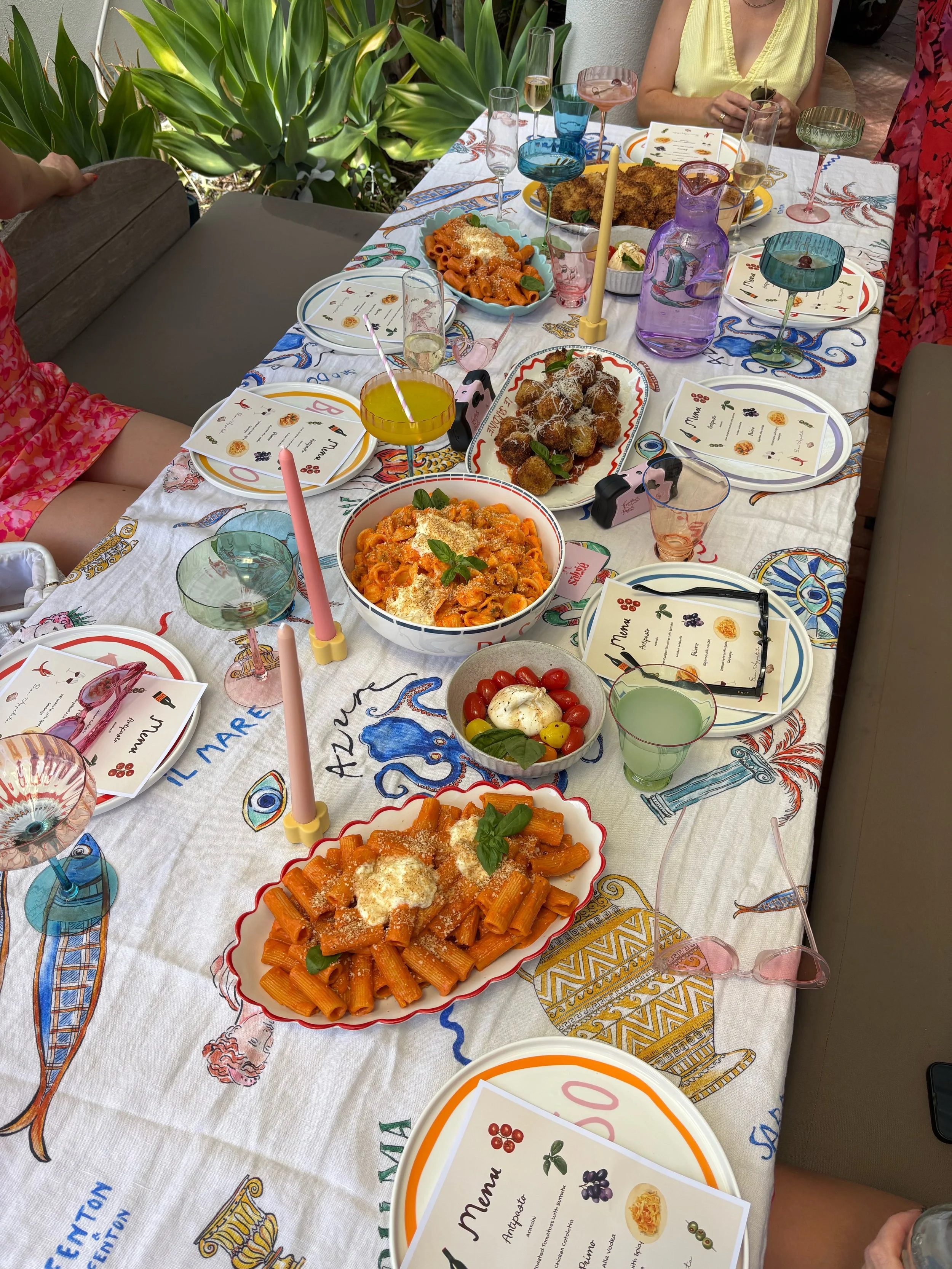 A colorful, festive Italian meal spread on a table with pasta dishes, salads, breaded fried items, drinks, and decorative candles, set outdoors with greenery in the background.