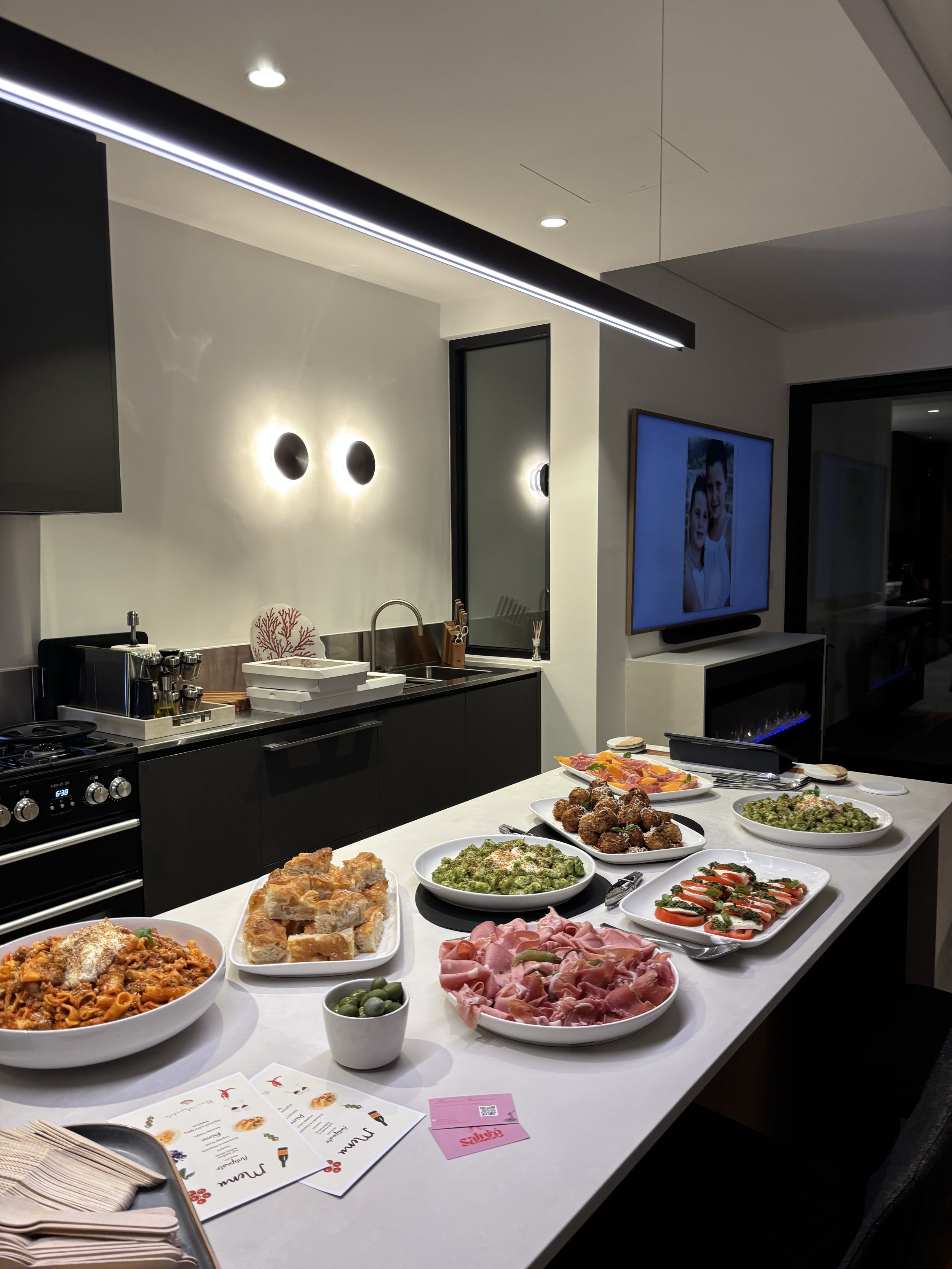 A kitchen island with various dishes including pasta, bread, olives, cold cuts, and vegetables, set for a meal in a modern kitchen with a TV on the wall.