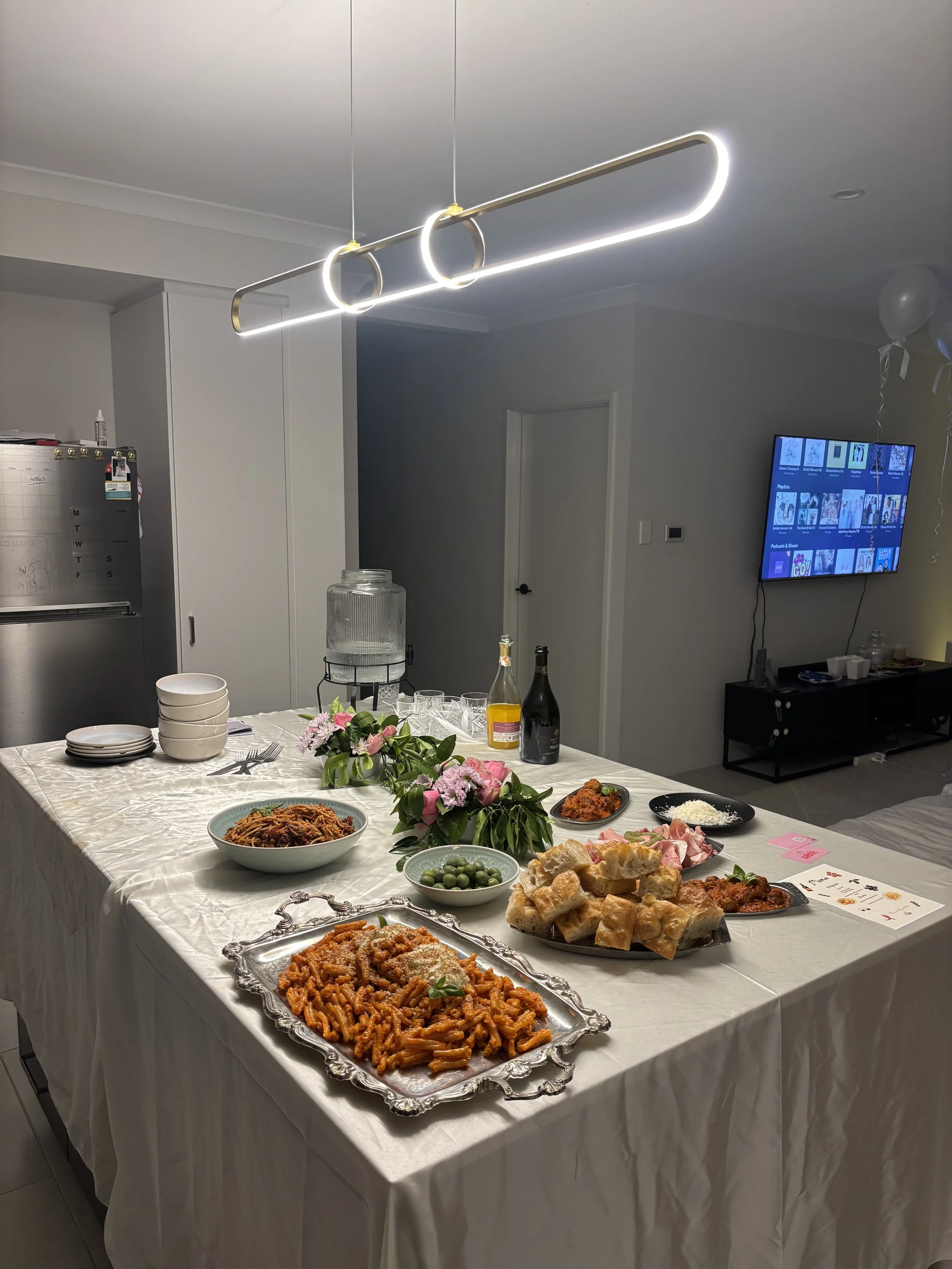 A dining table set for a celebration with pasta, bread, and appetizers, decorated with flowers, in a modern living space with a mounted TV.