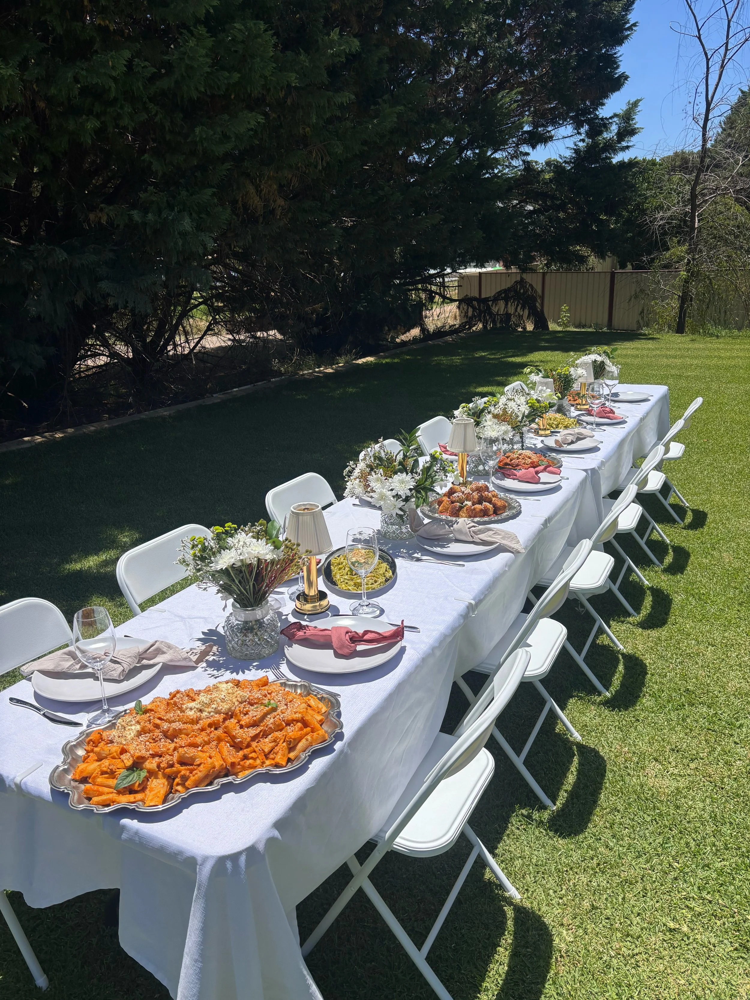 Outdoor dining table set for a meal with plates, glasses, floral centerpieces, candles, and various dishes, under trees on a sunny day.