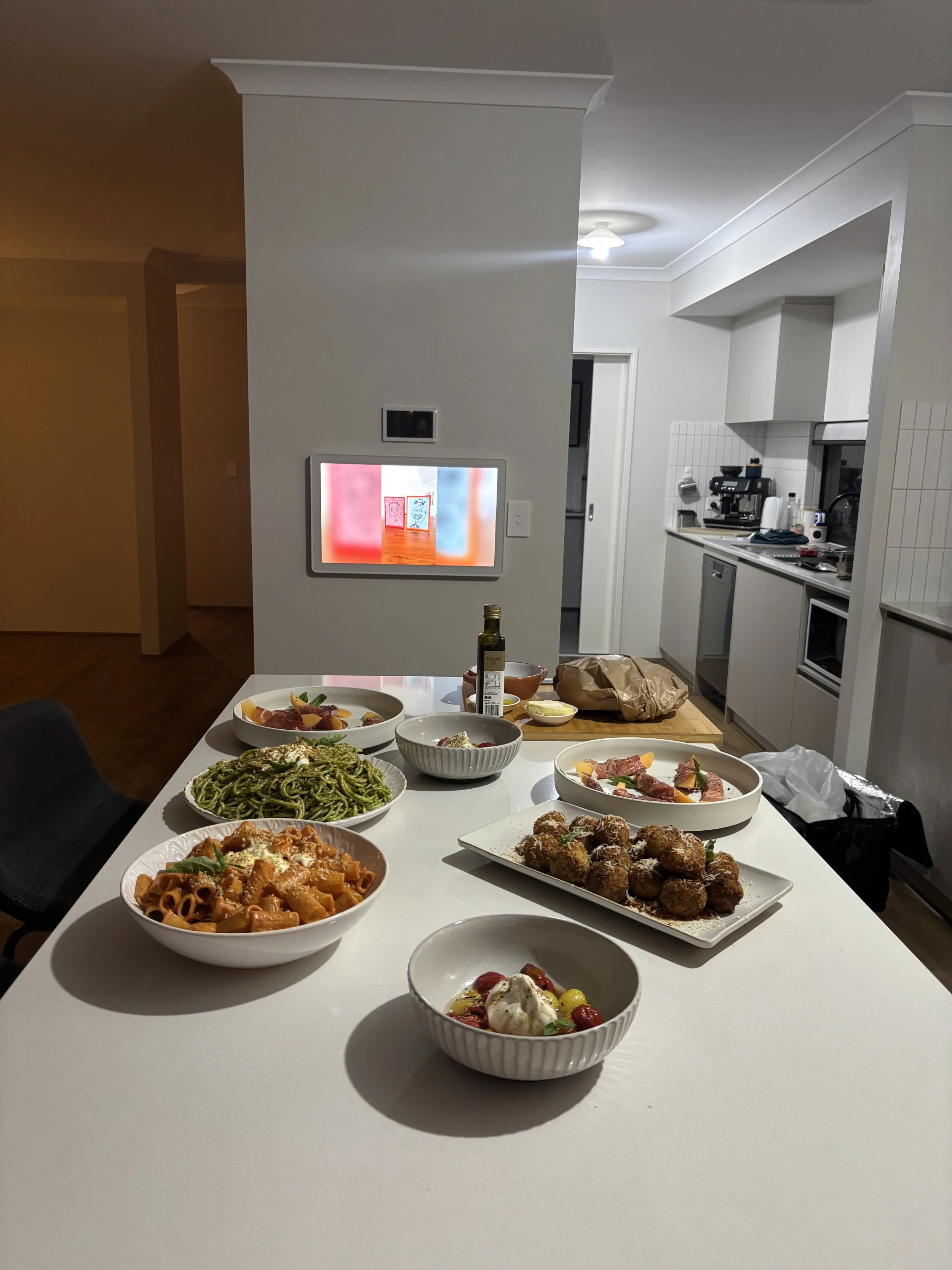 Dining table with various dishes including pasta, meatballs, and salad, set in a modern kitchen with white cabinets and appliances.
