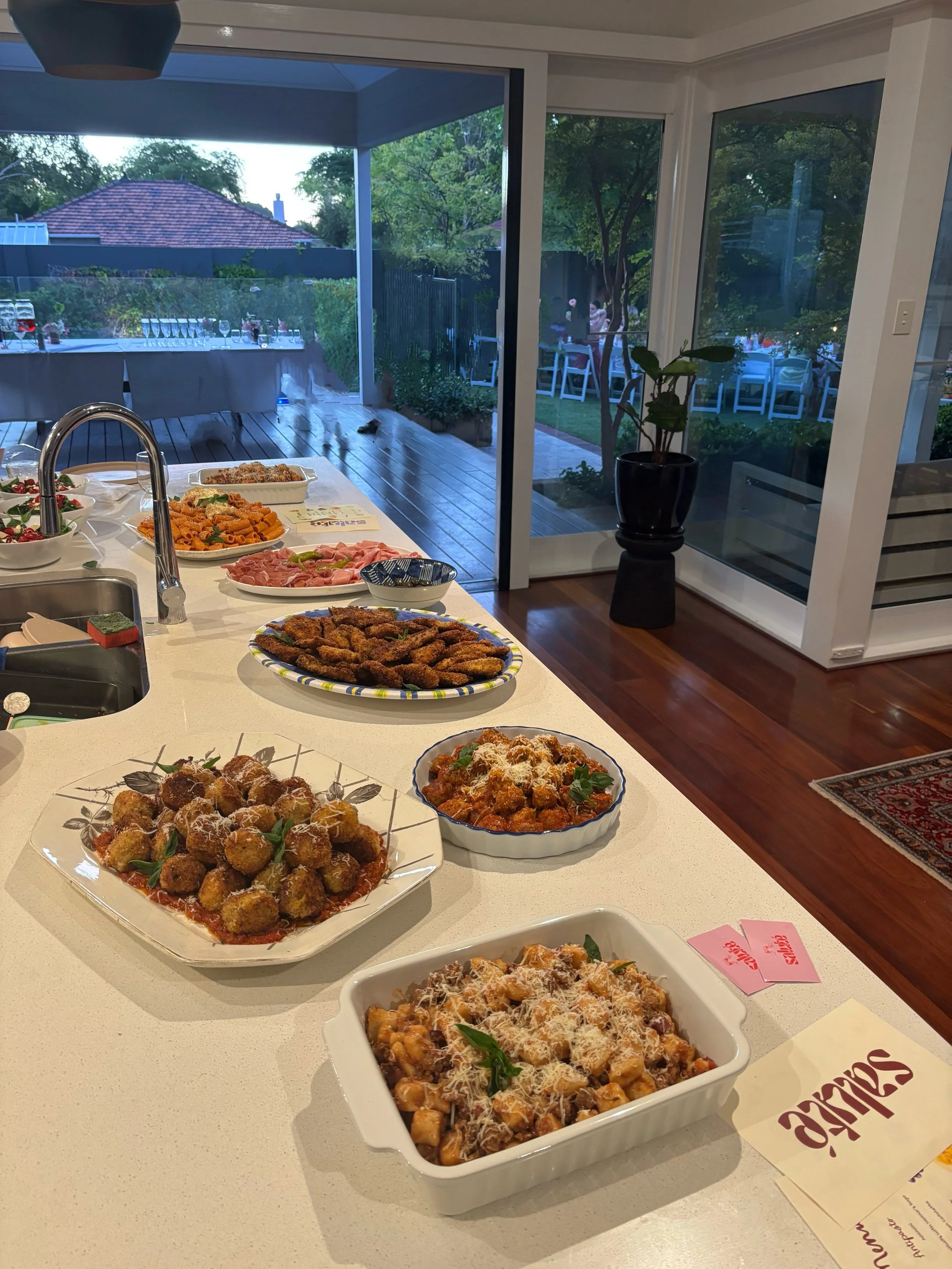 A kitchen island with various dishes of Italian food, including meatballs, pasta, and breaded chicken, with a sliding glass door leading to an outdoor patio with trees and chairs.