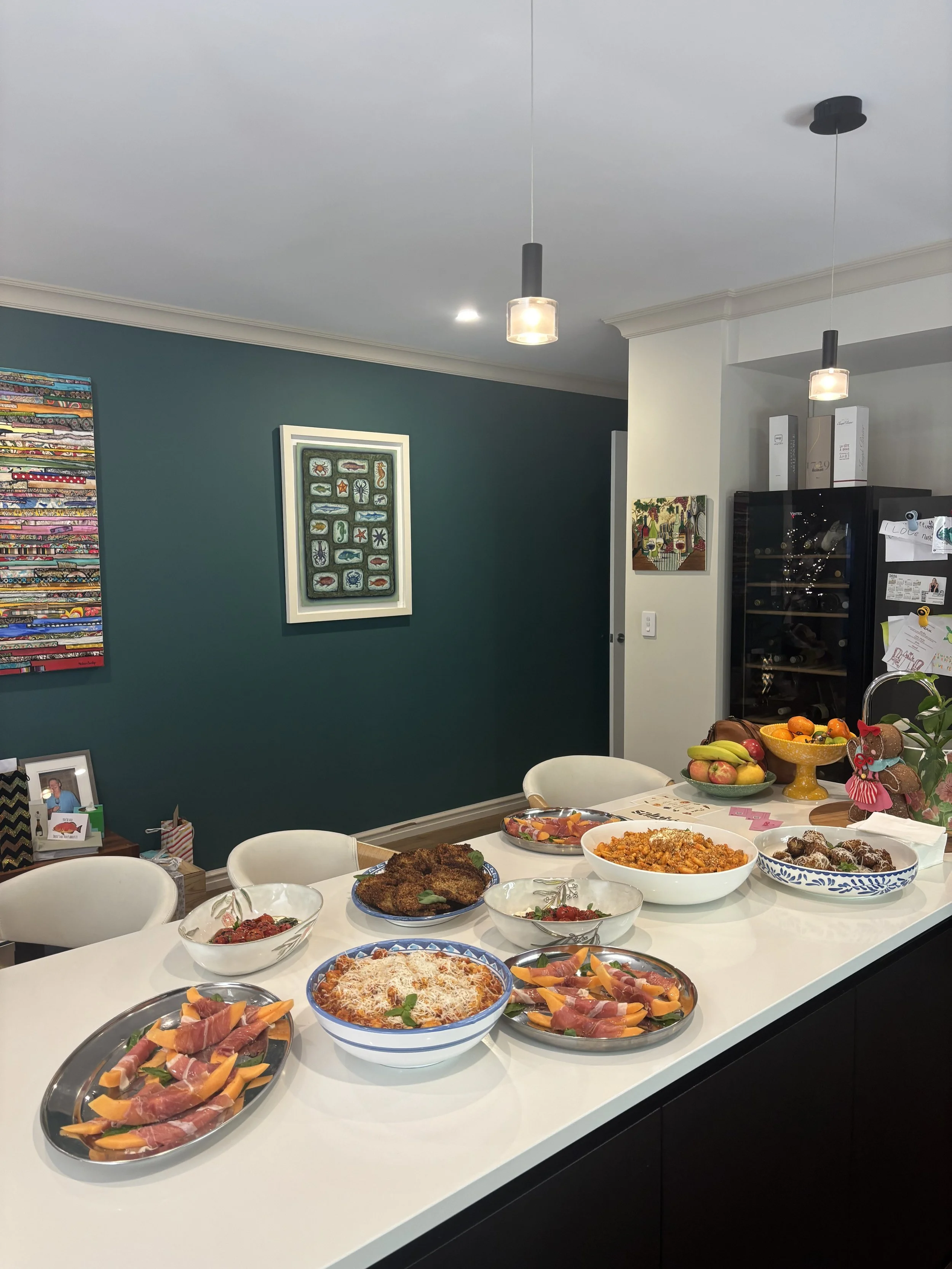 A kitchen island with various dishes and a bowl of fruit, with colorful artwork on the wall and pendant lights hanging from the ceiling.