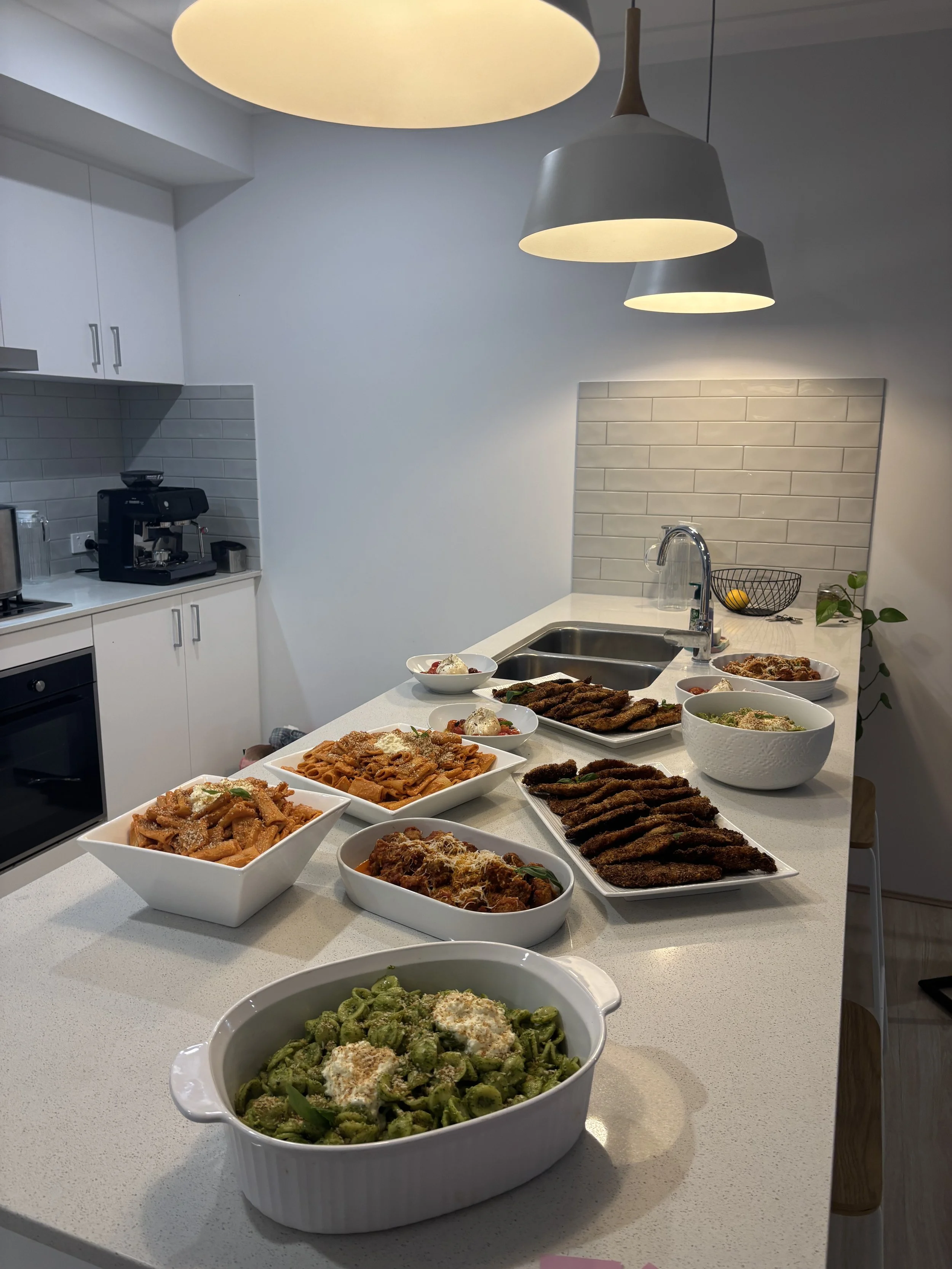 A kitchen counter with various bowls and plates of pasta, breaded meat, and salad, set for a meal, with modern lighting overhead.