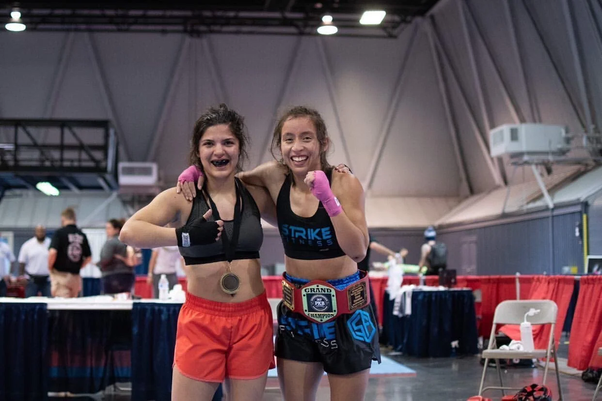 Dos luchadoras con ropa deportiva posan después de un combate, ambas sonrientes y luciendo cinturones de campeona. Una levanta el pulgar con la mano derecha, vistiendo pantalones cortos y un sujetador deportivo negro, con una medalla al cuello y vendas rosas. La otra luchadora, con pantalones cortos y un sujetador deportivo negro con detalles azules, rodea a la otra luchadora con el brazo izquierdo, sonriendo. Al fondo se ve un gimnasio con gente y equipo.