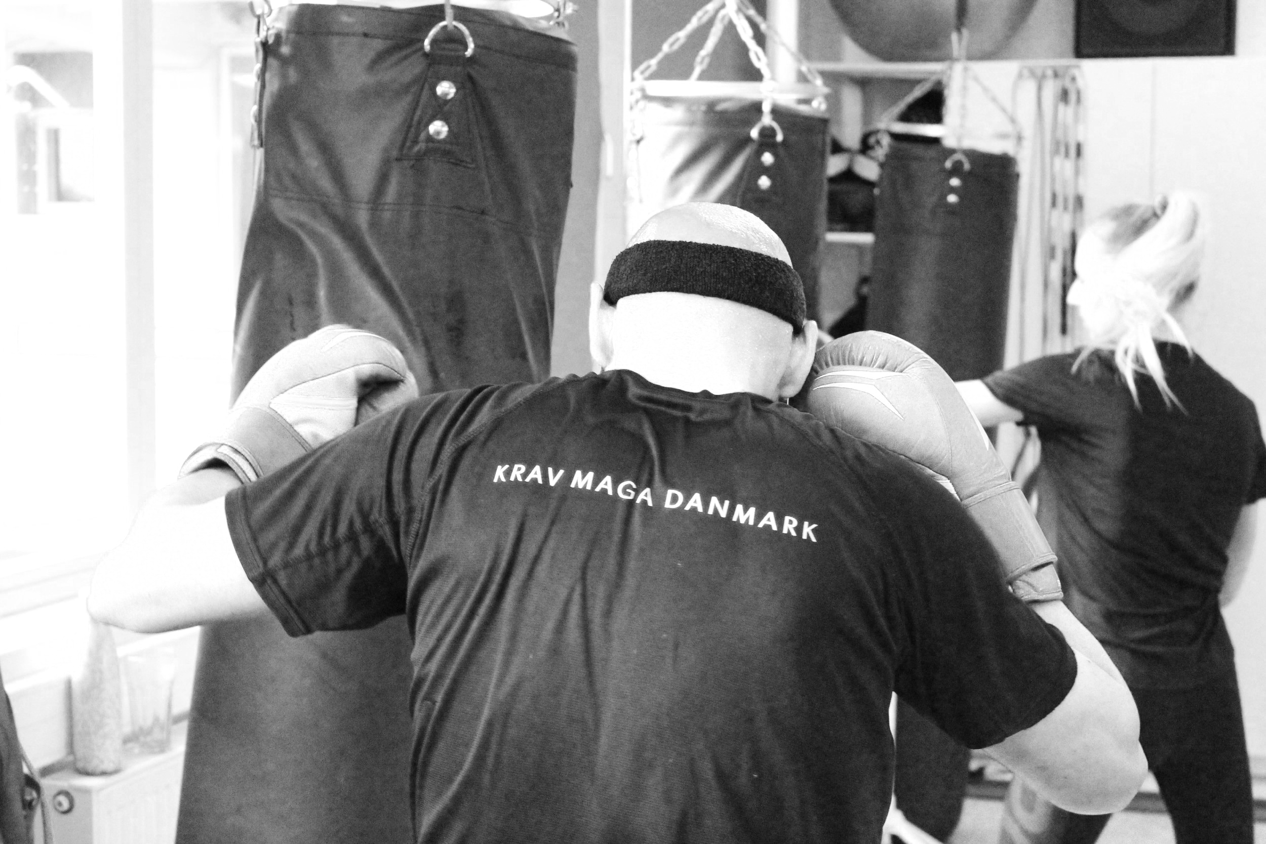 Person i sort t-shirt med teksten 'KRAV MAGE DANMARK' træner i kampsportslokale med sandsække og andre træningsudstyr.