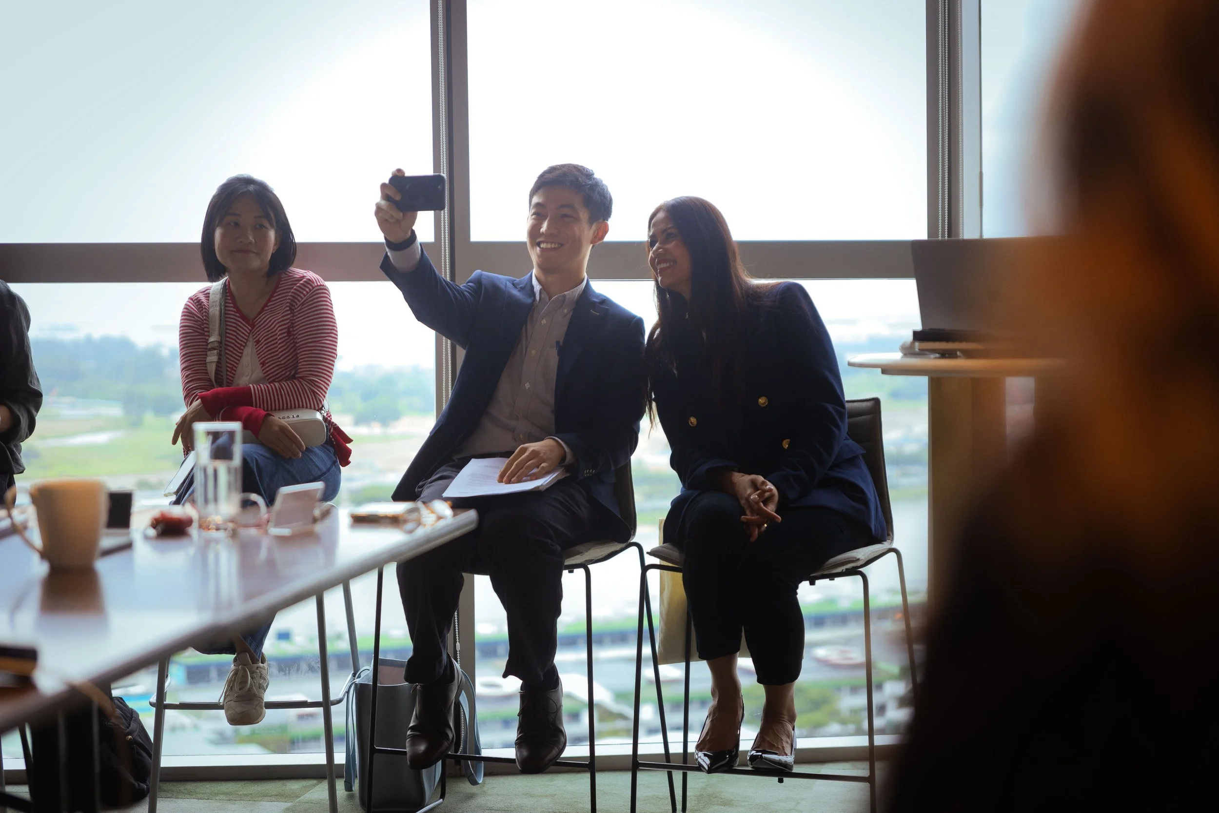 People sitting around a table in a conference room with large windows, one man taking a selfie with a smartphone while others look on.