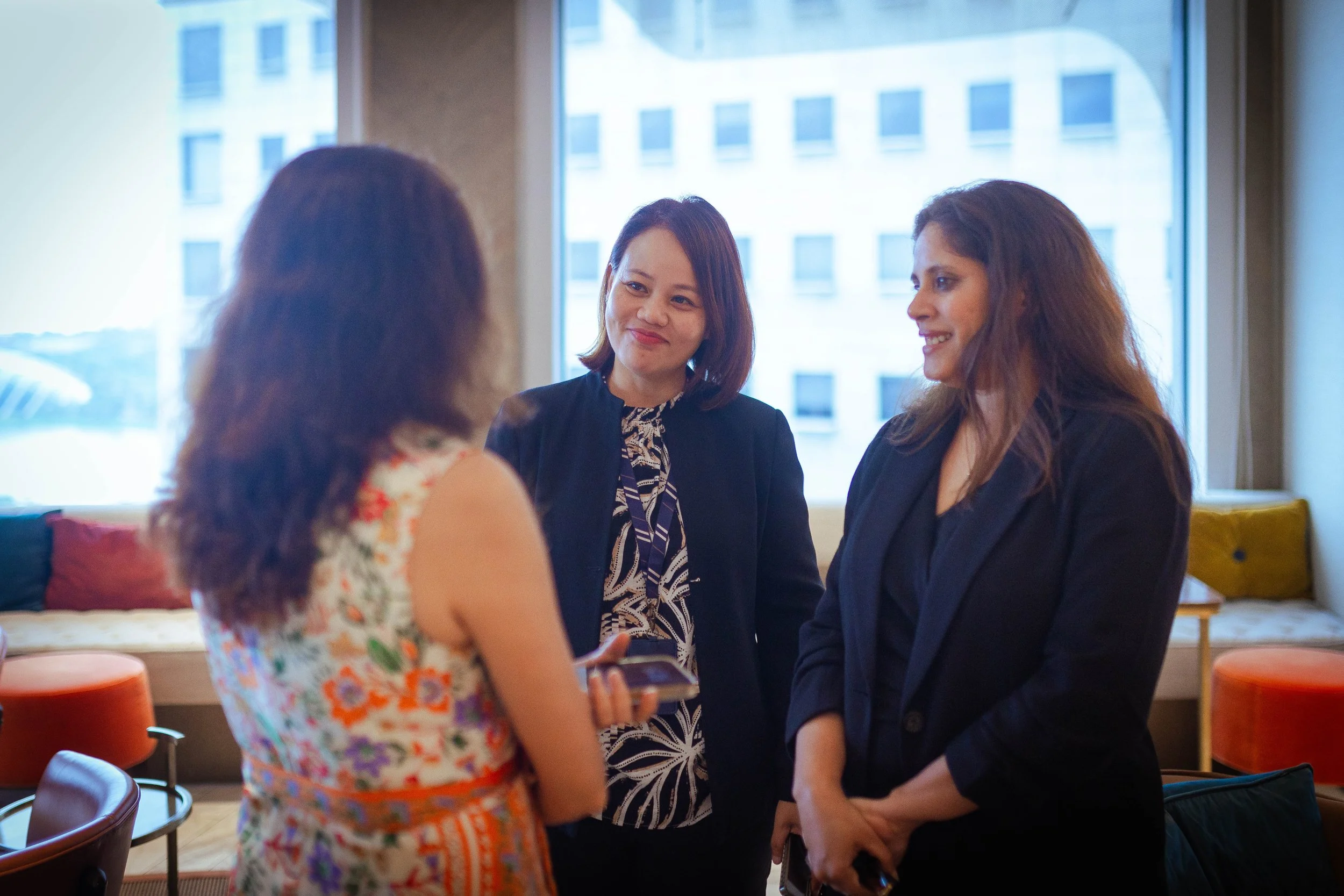 Three women engaged in conversation indoors, with a cityscape of tall buildings visible through the large window behind them.