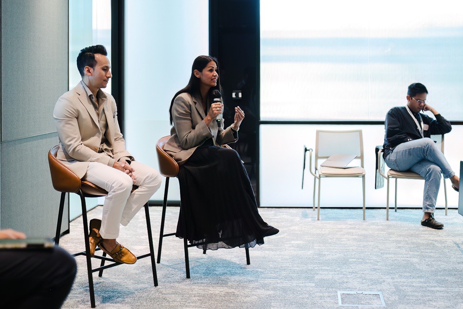 Three people seated in a panel discussion or conference room, with a woman holding a microphone and speaking, while the others listen; modern interior with chairs and large windows.