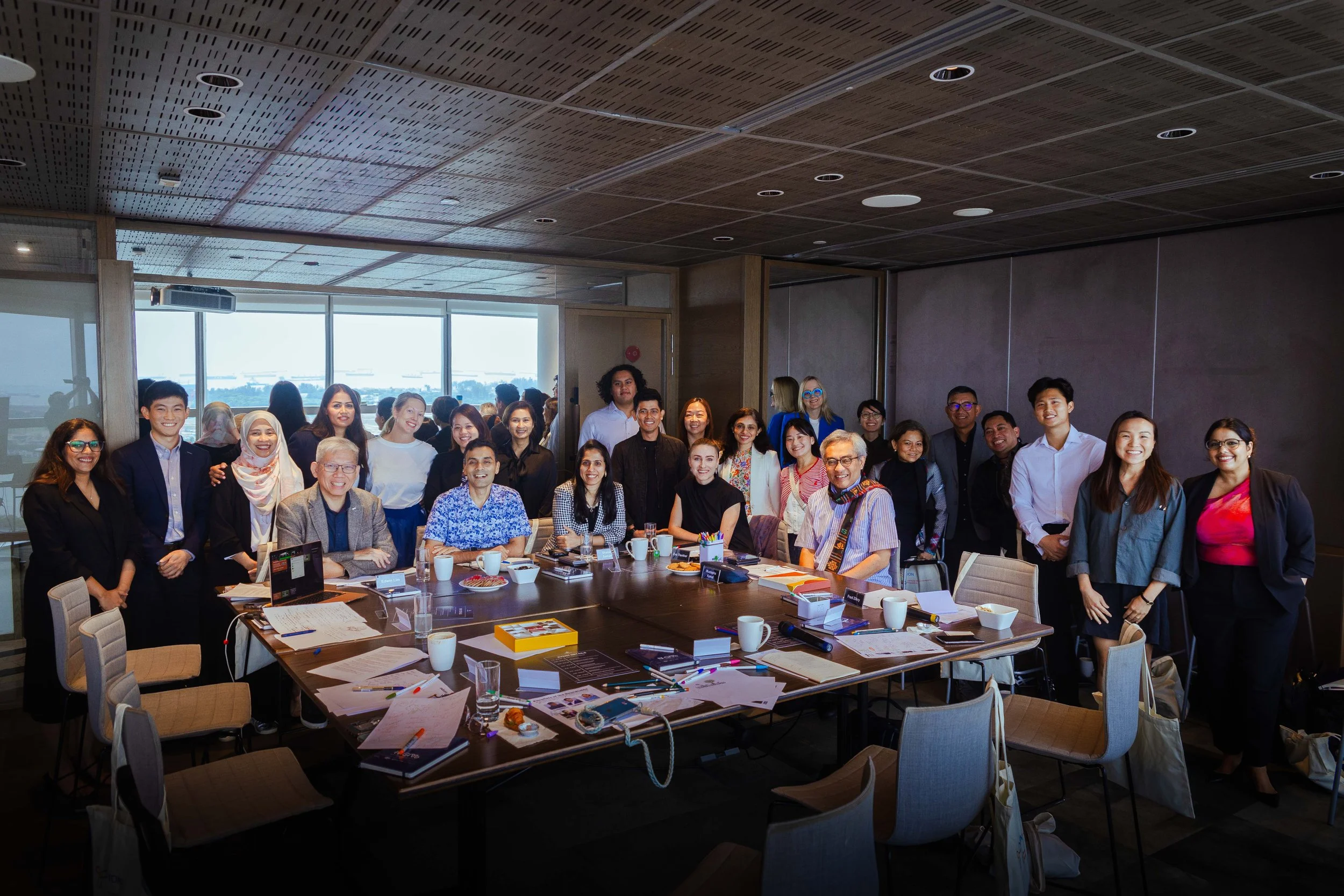 A diverse group of people gathered in a conference room for a meeting, with some seated around a large table and others standing behind. The table is cluttered with papers, notebooks, cups, and office supplies. The room has large windows letting in natural light.