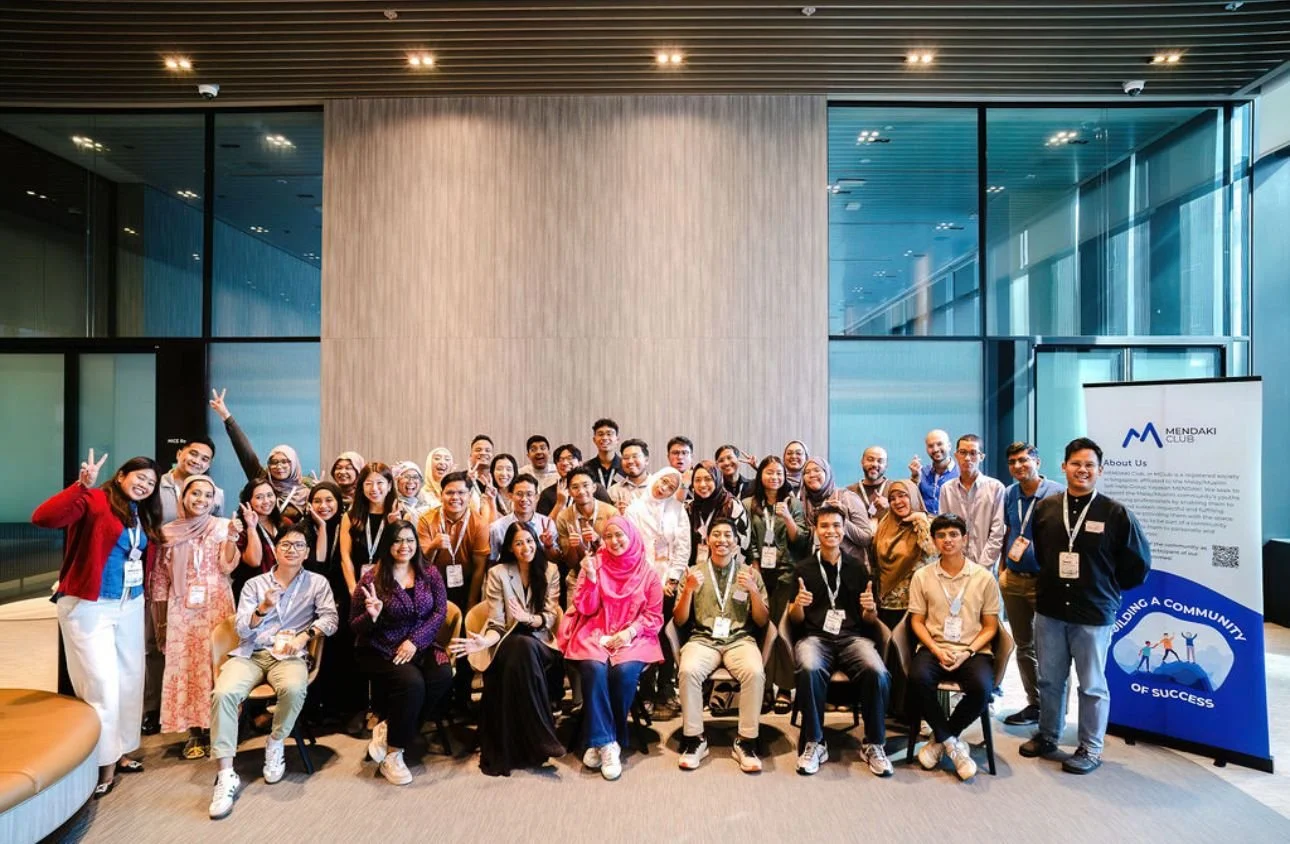 A large group of diverse people, including men and women of various ages and ethnicities, posing together indoors at a professional event or conference. They are smiling, some making peace signs, and wearing conference badges.