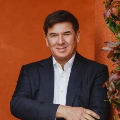 A man in a suit standing in front of an orange wall with a plant on the right side.
