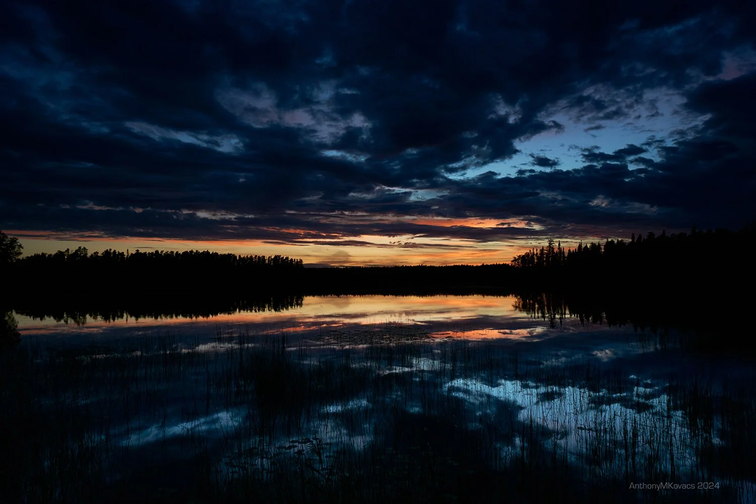 A twilight scene over a lake with dark clouds in the sky and trees along the horizon, with a colorful sunset reflected in the water.