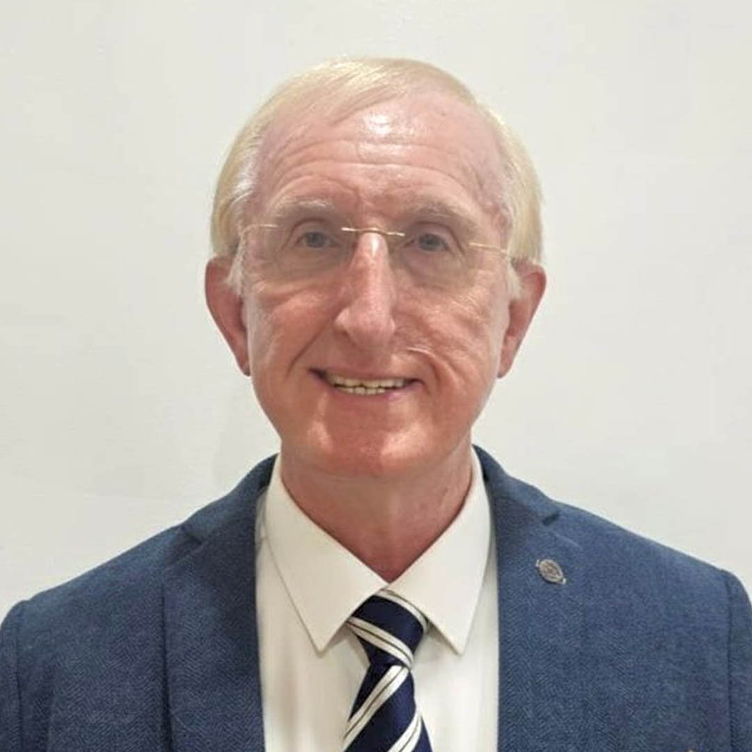 A middle-aged man with glasses, light blonde hair, wearing a blue blazer, white shirt, and striped tie, smiling in front of a plain neutral background.