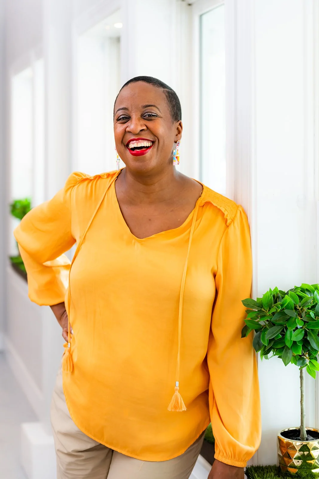 A woman with a short haircut wearing a yellow blouse and smiling in a brightly lit room with white walls and a potted green plant.