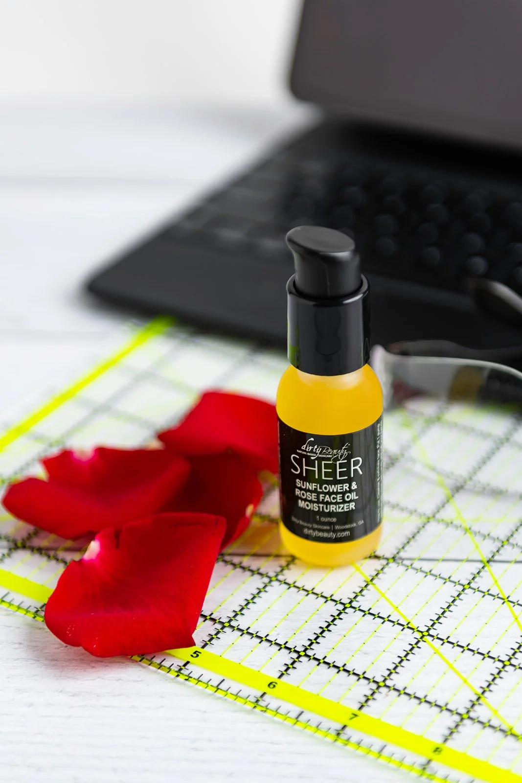 A small bottle of 'Sheer Sunflower & Rose Face Oil Moisturizer' on a cutting mat with red rose petals and a blurred laptop in the background.