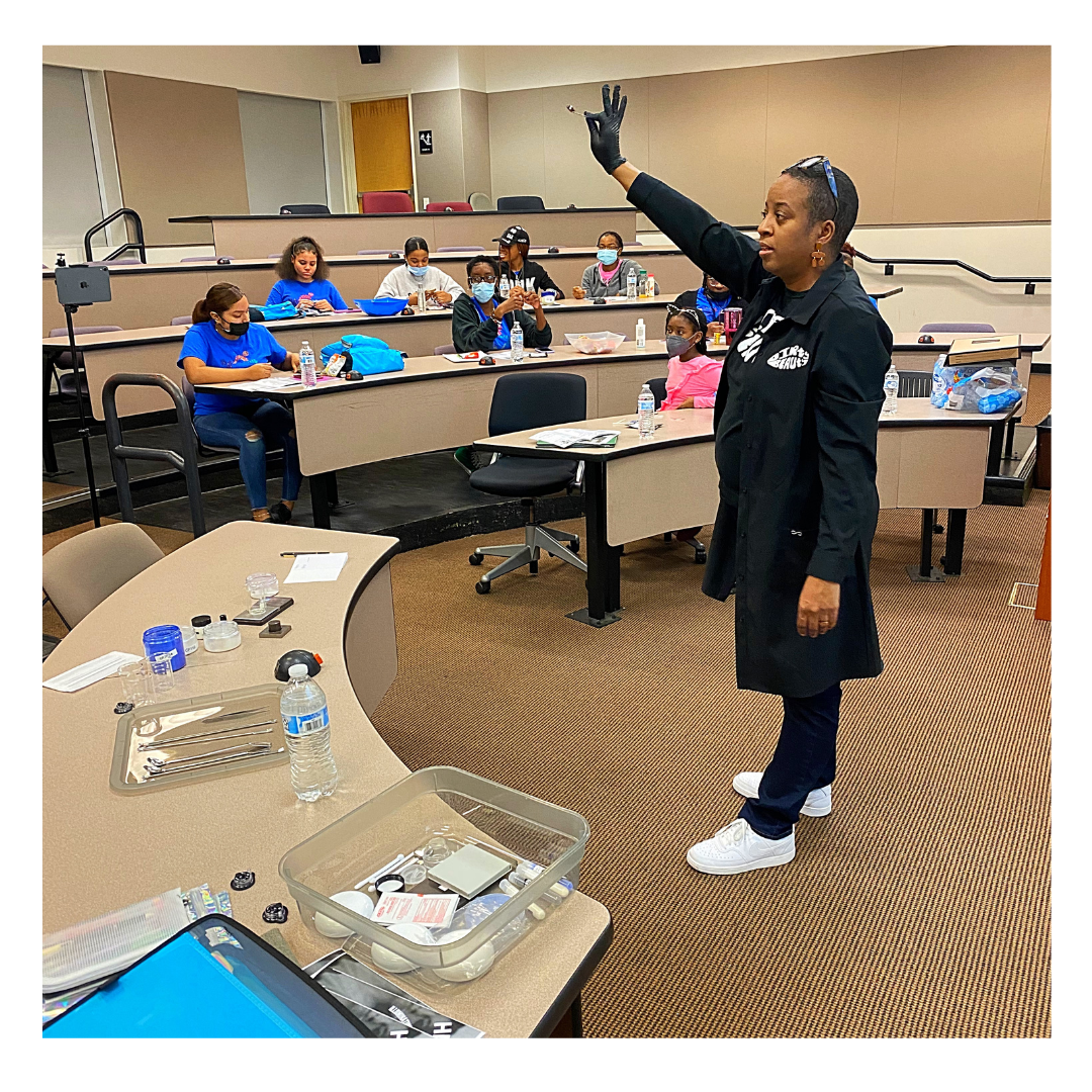 Instructor standing in front of a classroom of children sitting at desks, raising her hand, wearing gloves, and wearing a black coat.
