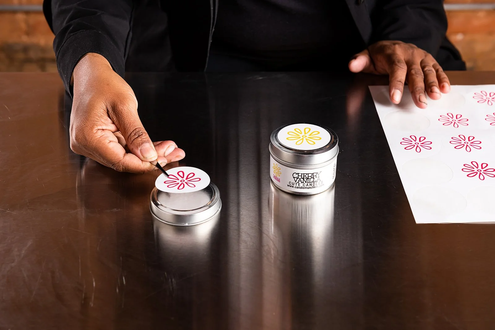 Person decorating candles with floral peel-off stickers, some stickers and a container of candle wax on a dark wooden table.