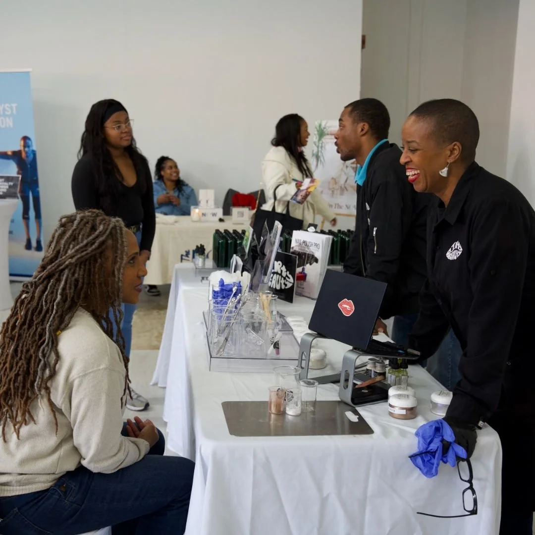 Two women at a booth talking to a woman sitting down at a table with products displayed. The women are smiling and engaged in conversation at an indoor event or market.
