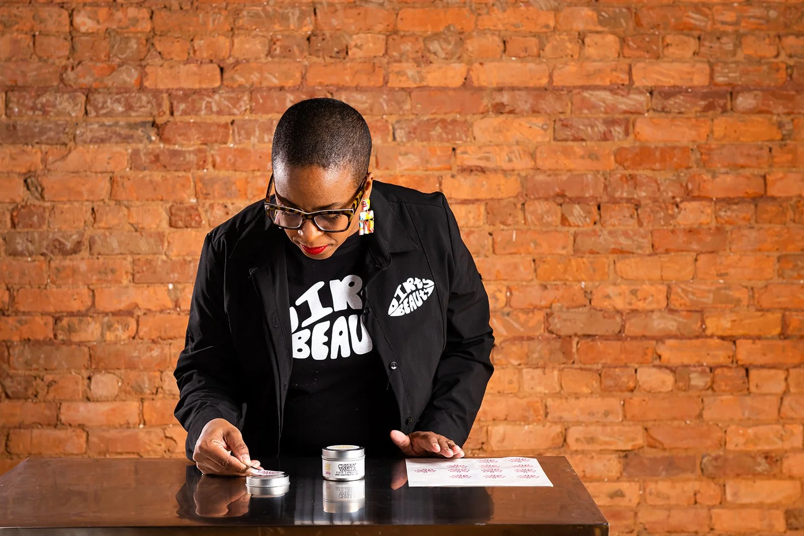 A person with short hair, glasses, and red lipstick examining small containers on a dark wooden table against a brick wall.