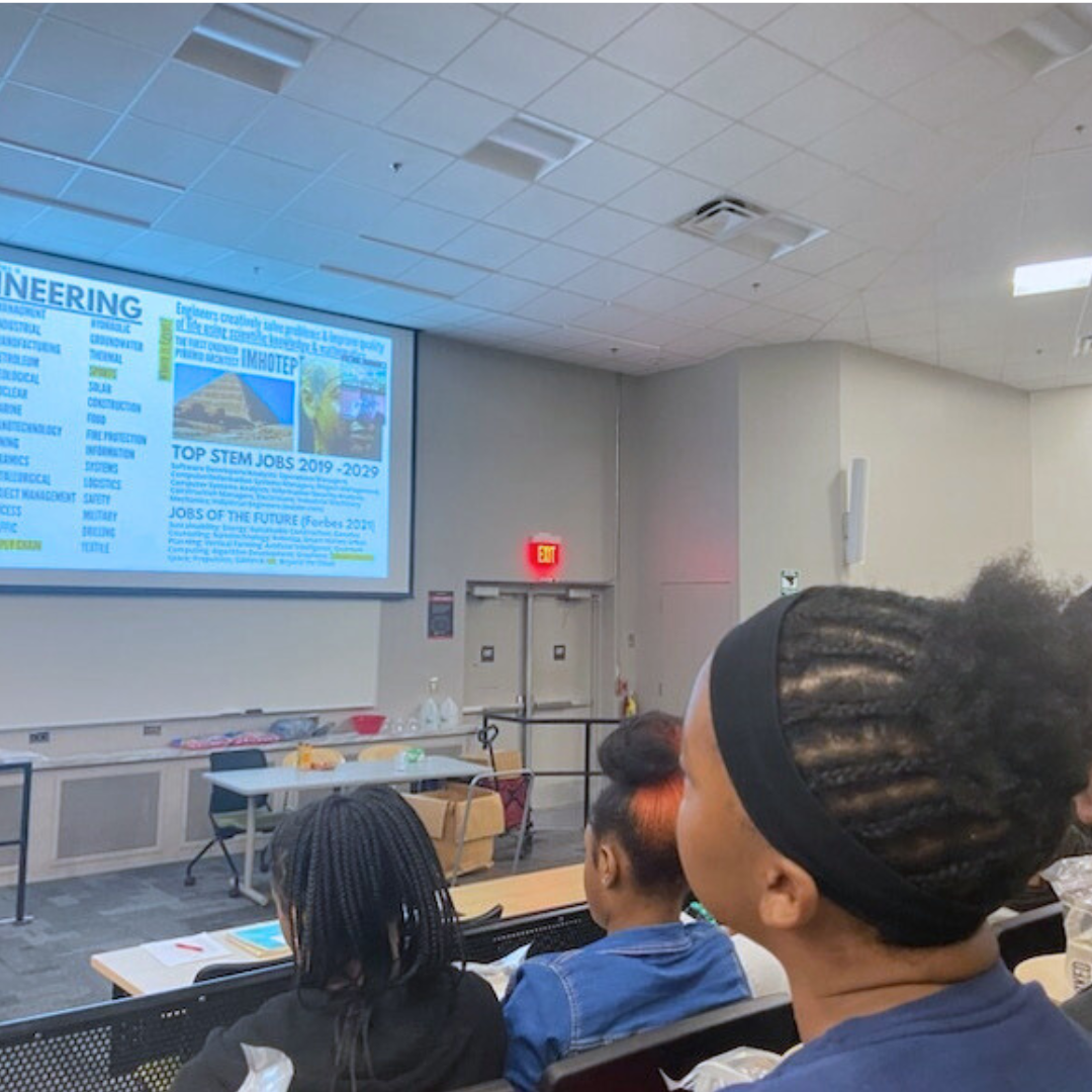 A classroom or seminar room with students watching a presentation on a large screen at the front of the room. The presentation slide contains text and images related to engineering and STEM careers.