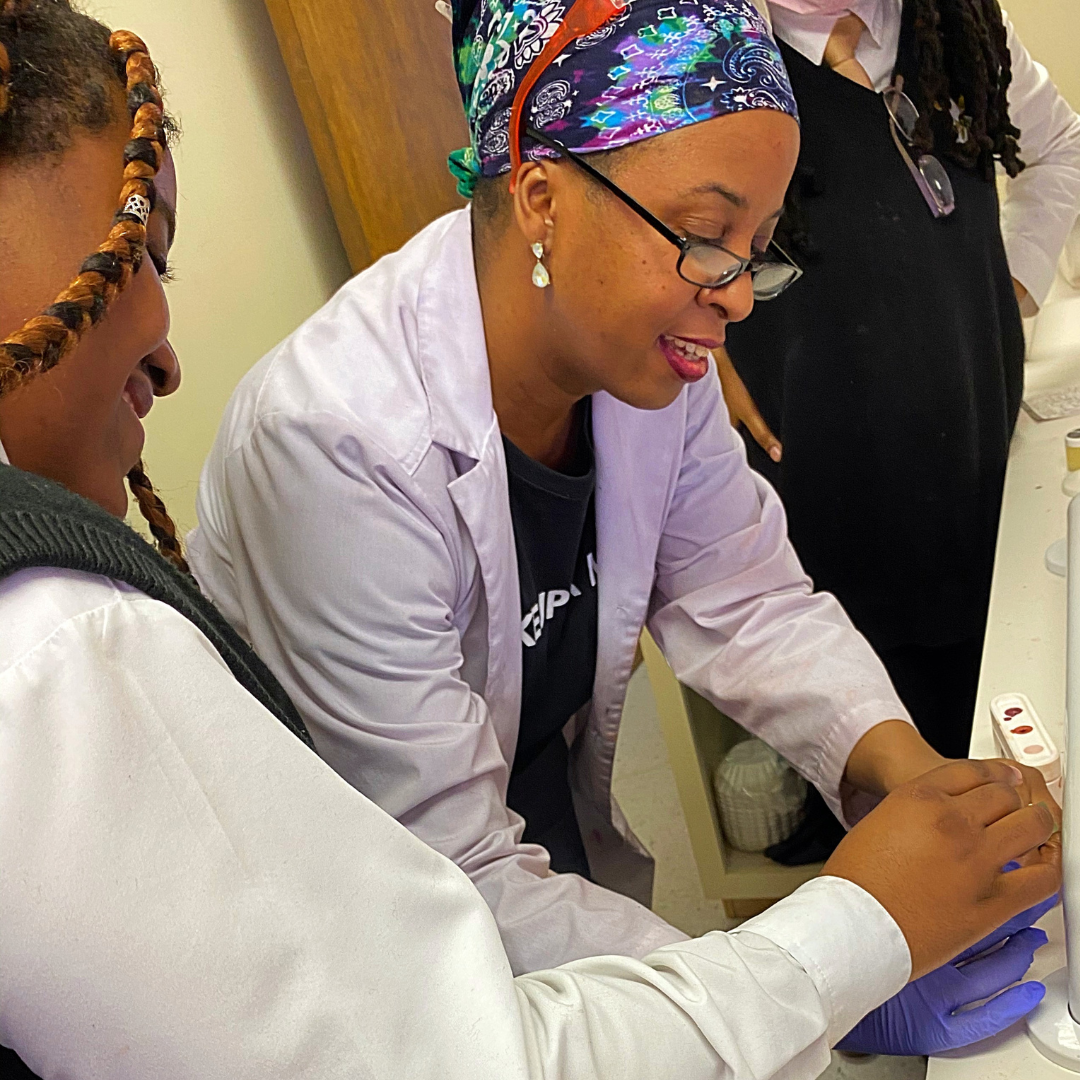 A woman wearing a colorful headscarf, glasses, and a lab coat, is assisting another person in a lab setting with an experiment, with laboratory equipment visible on the table.