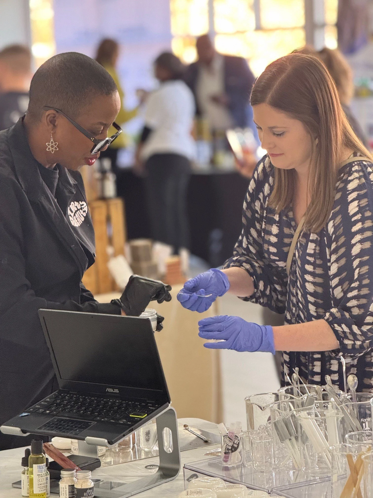 Two women in a laboratory or workshop setting, wearing gloves, working with scientific tools and equipment, with a laptop on the table in front of them.