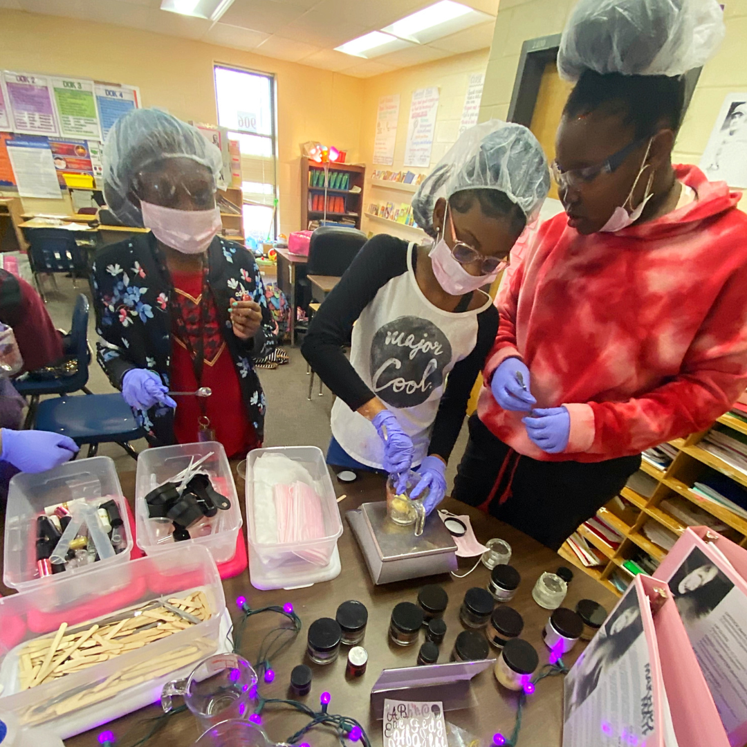 Three girls in a classroom, wearing hair nets, face masks, and gloves, are conducting a science experiment with various jars and materials on a table.